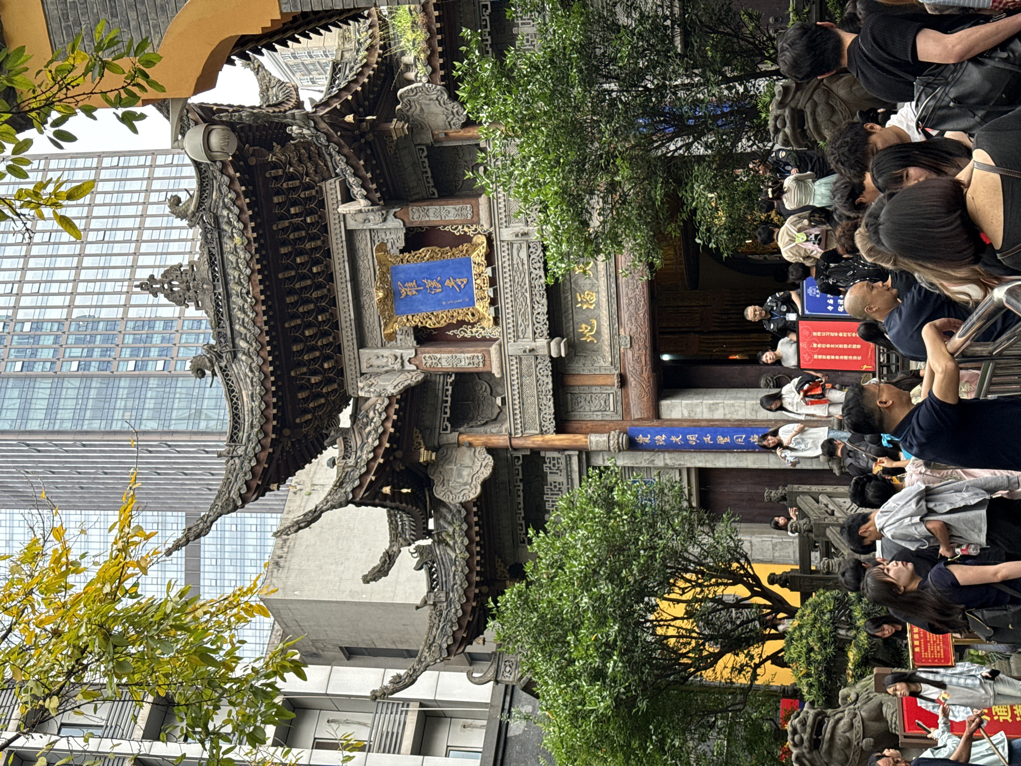 Here's a description of the image:
The image captures an urban scene featuring a traditional Chinese temple facade juxtaposed against modern skyscrapers. The architecture of the temple is ornate, with layered, curved roofs and intricate carvings. A blue and gold sign with Chinese characters is prominently displayed.
A crowd of people are seen gathered near the entrance, possibly waiting to enter the temple. They appear to be a mix of ages and are dressed in casual clothing. A metal barrier and a uniformed security guard are present, suggesting crowd management measures.
The background is dominated by tall, contemporary buildings with many windows, contrasting with the traditional style of the temple. A few trees with green foliage provide some natural elements in the scene.
The image conveys a sense of cultural harmony between ancient tradition and modern city life. It is likely located in the city of Yuzhong District, China, given the cultural details.