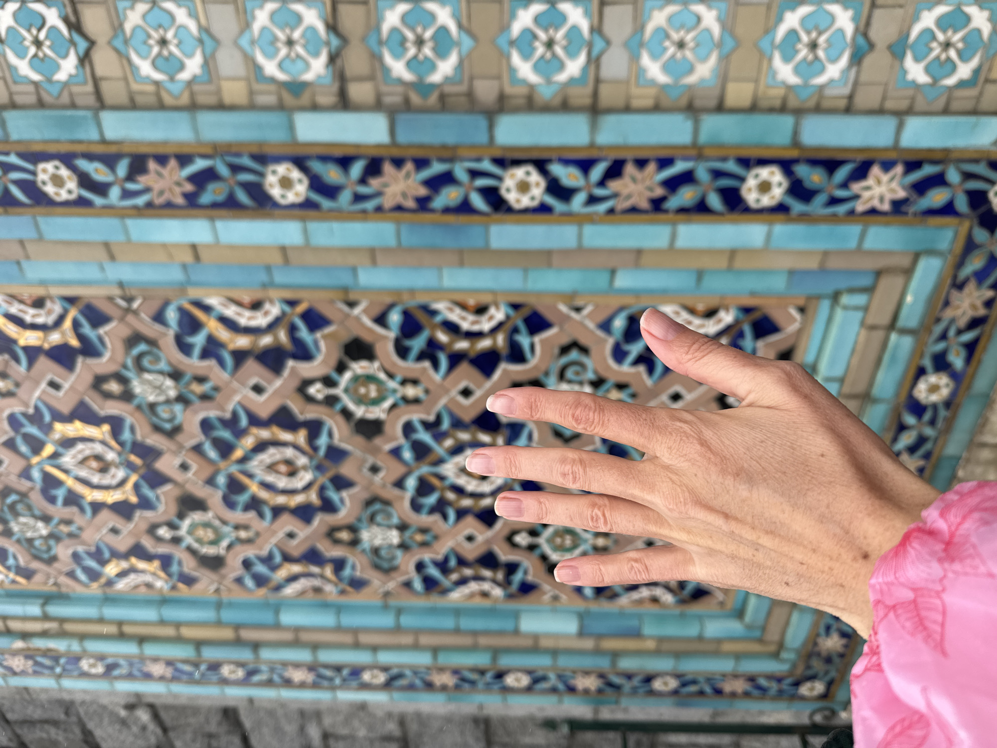 A hand extends toward the ceiling of the Peterhof Palace in Saint Petersburg, Russia. The ceiling is ornate, tiled in a pattern of repeating geometric shapes in blue, white, beige, and gold. The hand is light-skinned with short fingernails and is angled slightly to the left. Part of a pink sleeve is visible at the right edge of the image. The background is slightly out of focus.