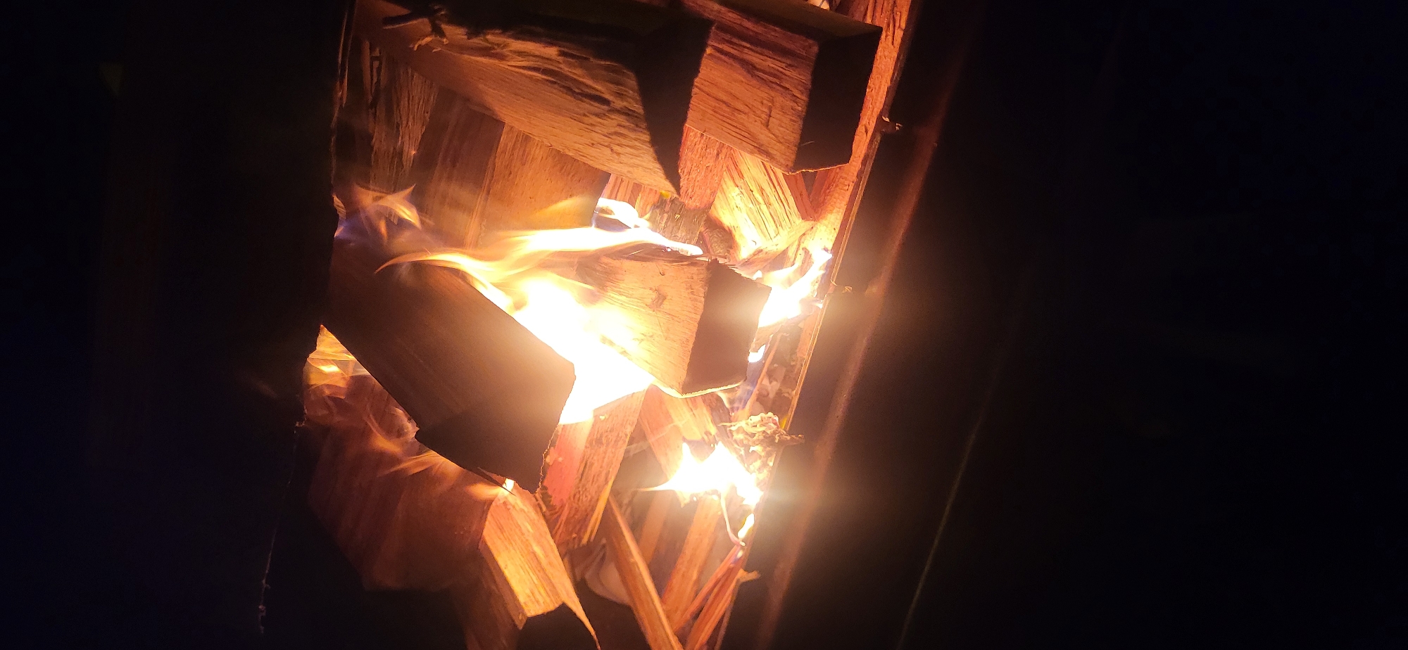 The image shows a close-up view of a fire pit filled with burning logs. The logs are stacked and appear to be hardwood, with flames of various colors, primarily yellow and orange, emanating from them. The fire is intense and casts a warm glow on the surrounding wood. The background is dark, suggesting nighttime or a dimly lit environment. The fire pit itself appears to be made of metal and has a dark, matte finish. There are no people or other objects visible in the image.