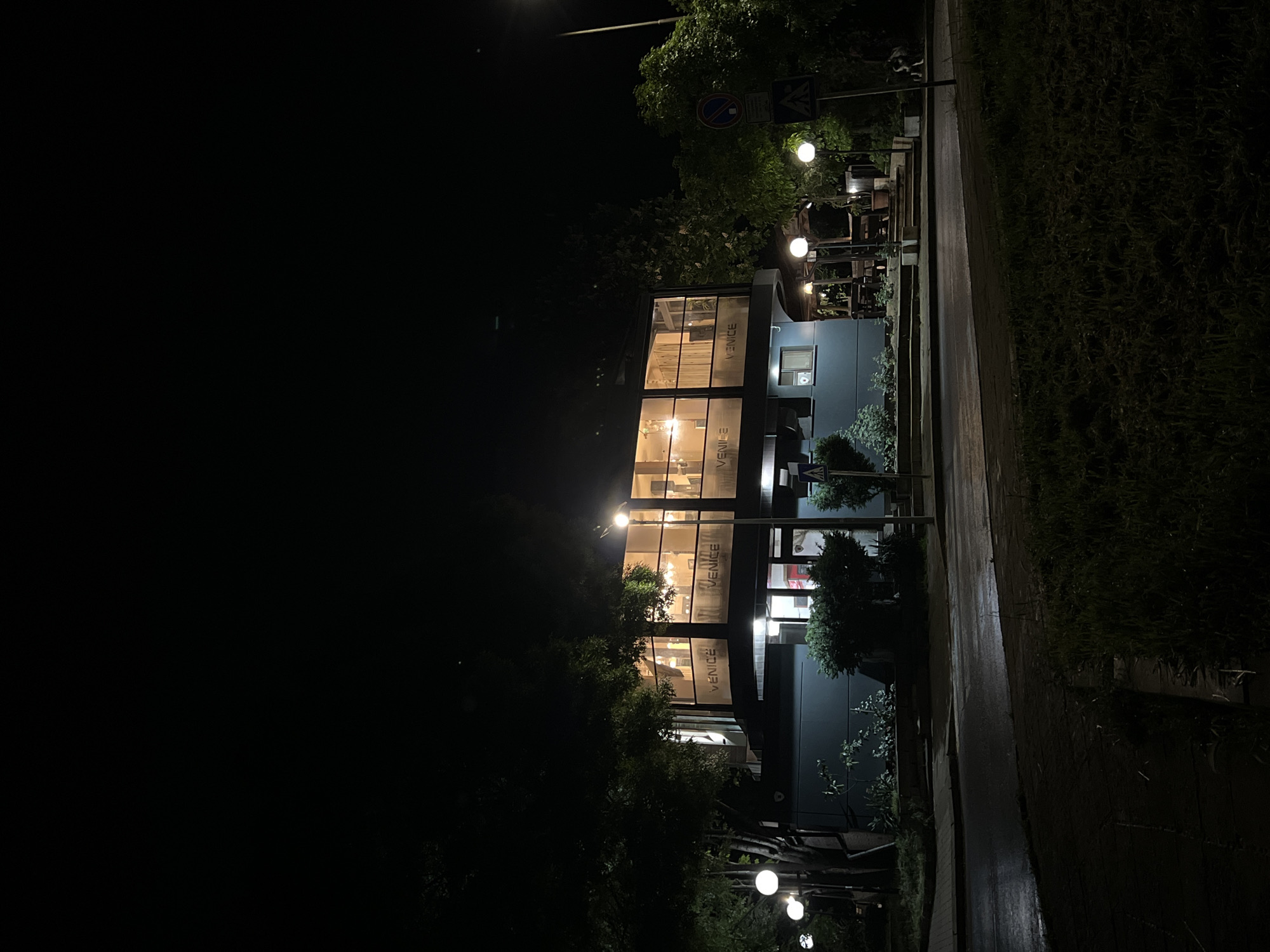 The image depicts a nighttime scene in Burgas, Bulgaria. A two-story building with large glass windows is illuminated from within, displaying the word "VENICE" vertically on the windows. The building is situated on the edge of a street, with a sidewalk and adjacent vegetation on the right. Streetlights provide additional illumination along the sidewalk. Trees surround the building and obscure part of the facade. No people are visible in the image. A signpost with traffic signs is present near the building. The street is dark and wet.