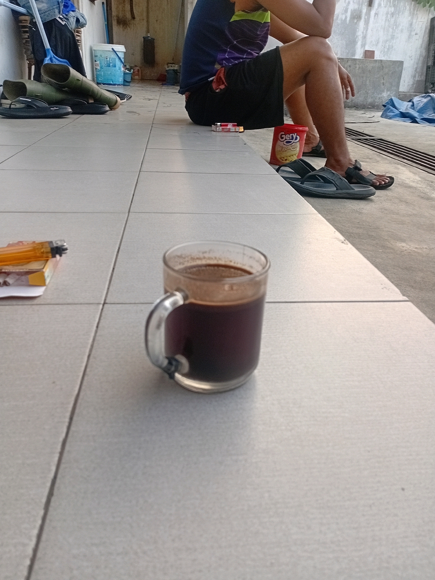A person is sitting on a raised concrete step with their legs extended in front of them, wearing black shorts and a dark blue t-shirt with a purple and green design. In front of the person, on the tiled ground, sits a glass mug filled with dark liquid, presumably coffee, and a cigarette pack with a lighter on top. To the right of the mug, a red plastic container is visible. The person is wearing dark flip-flops on their feet, and another pair of dark flip-flops is placed on the tiled ground near a pair of green rubber boots. The scene appears to be outdoors in a semi-urban environment, possibly a residential yard or an open workshop area, characterized by concrete flooring, a simple building in the background, and some tarpaulin sheets. The lighting suggests it is late afternoon or early evening. The location is identified as Tangerang, Indonesia.