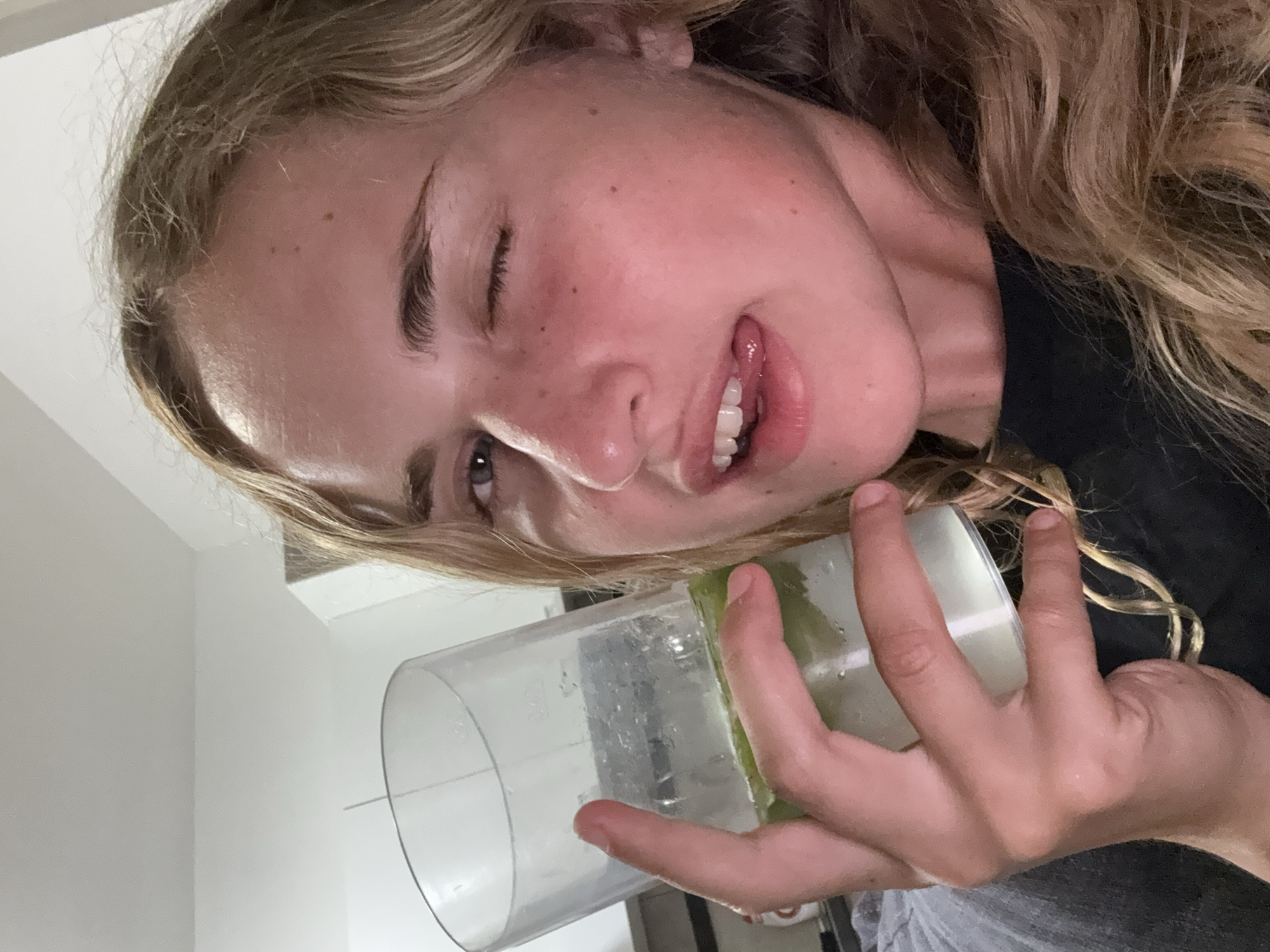 A young woman with blonde, wavy hair and fair skin is holding a clear plastic cup containing a green liquid and ice. She is looking towards the camera and winking, with her tongue slightly sticking out of her mouth. She is wearing a black t-shirt. The background is blurry but appears to be an indoor setting with white walls and a ceiling. The image was taken in Pula, Croatia.