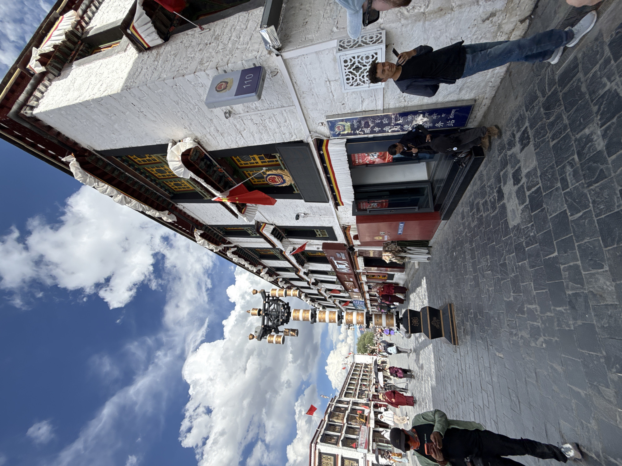 This vibrant photograph captures a bustling street scene in what appears to be a Tibetan cultural area, likely Chengguan District in China, under a bright, partly cloudy sky. The street is paved with dark stone blocks, and traditional multi-story buildings with white-washed walls and ornate dark wooden eaves line both sides.
The architecture features decorative window frames and numerous prayer flags in white, red, and yellow, adding to the cultural ambiance. Many buildings also display colorful traditional motifs in red, yellow, and black under their eaves. A red flag, possibly the Chinese national flag, hangs from one building. A prominent sign with the number "110" is visible on one of the buildings, suggesting it might be a public service or address marker. Other signs with Chinese characters are present but mostly unreadable from the distance.
Numerous people are seen walking along the street. Their attire varies, with some individuals in modern casual clothing like jeans and jackets, while others wear traditional dark red monastic robes, indicating a mix of locals, tourists, and possibly religious practitioners. In the foreground, a man in a green jacket and dark pants is seen looking at his phone. Another man in a dark jacket and blue jeans is also visible further down the street, holding a phone.
A distinctive feature of the scene is a series of large, golden prayer wheels mounted on tall pillars in the mid-ground, serving as a focal point and strongly suggesting the spiritual significance of the location. These prayer wheels are interspersed with ornate dark street furniture. The bright daylight and clear blue sky with scattered white clouds indicate a pleasant day. The overall atmosphere is one of active urban life steeped in rich cultural heritage.