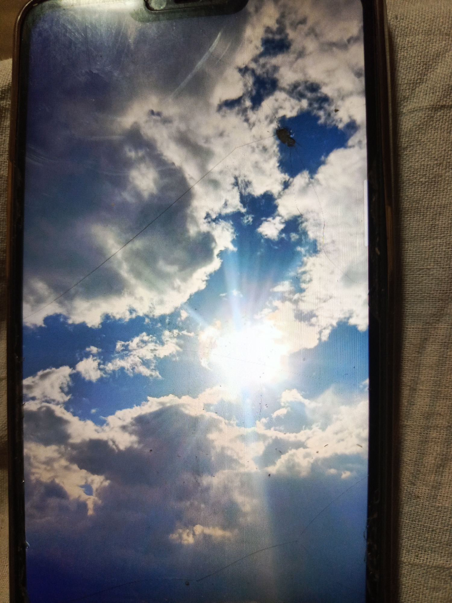 The image is a photograph of a smartphone screen displaying a bright sky with scattered clouds and the sun. There are visible scratches and smudges on the screen. The background fabric suggests the phone is resting on a textured surface. The scene does not provide any information or context related to the city of Svoboda, Russia.