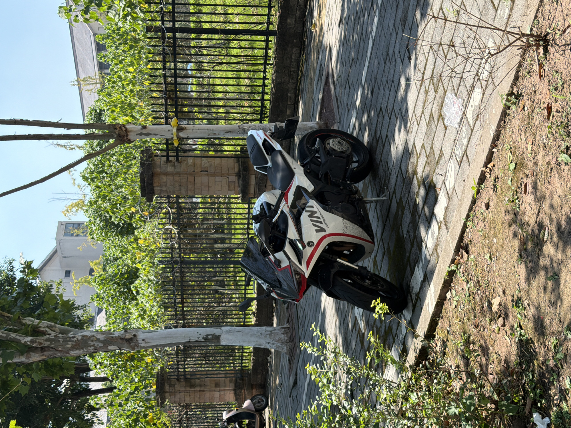A white and red sport motorcycle is parked on a paved sidewalk next to a black metal fence. The motorcycle has the word "VINY" printed on its side. To the left of the motorcycle, a light-colored scooter is partially visible. Behind the fence, lush green foliage and trees are abundant, with buildings visible in the background. The ground in front of the sidewalk is covered in dirt and sparse vegetation. The scene appears to be a sunny day. The location is identified as Jiangjin District, Chongqing, China.