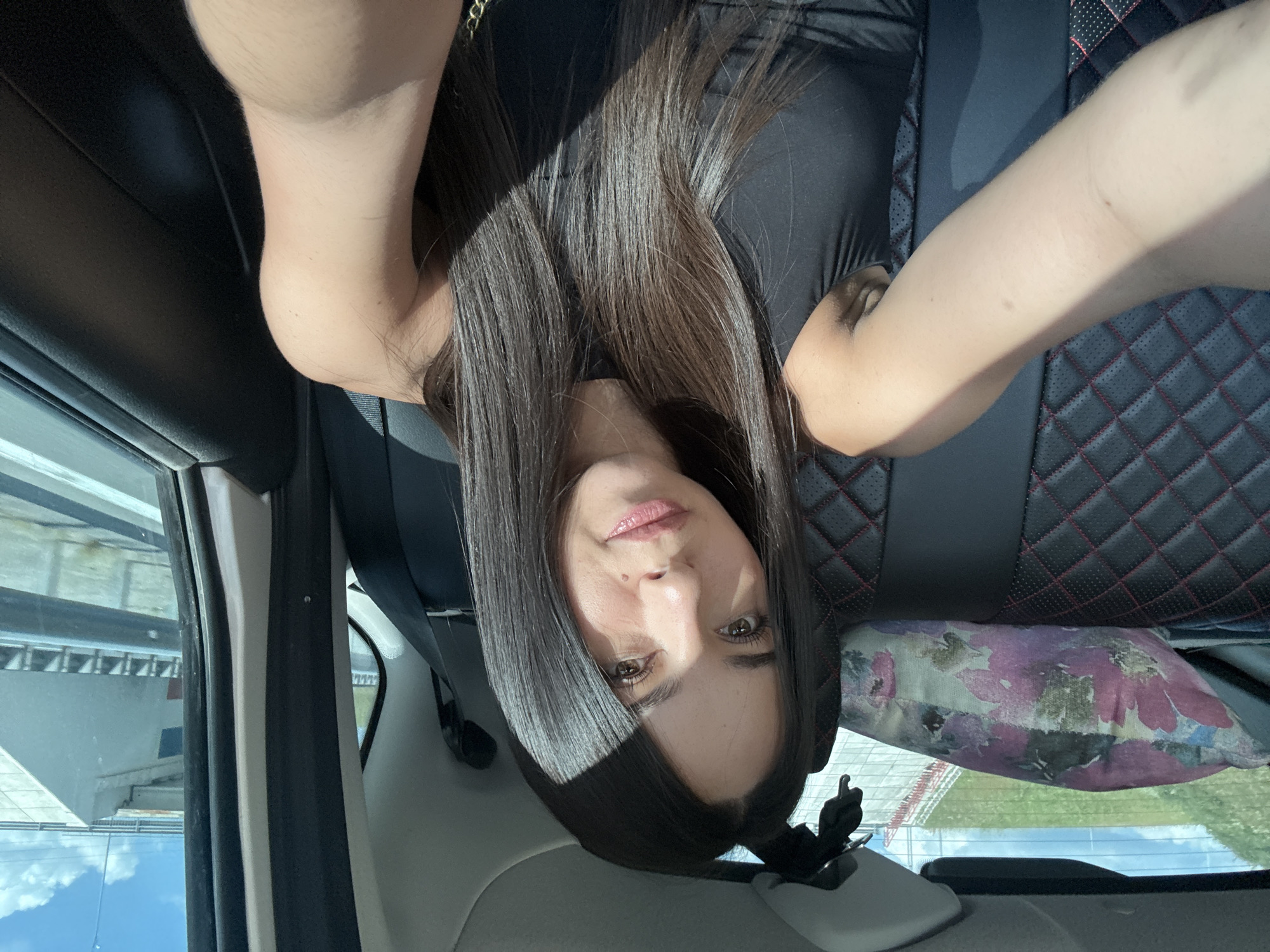 A woman with long, dark hair is sitting in the back seat of a car, taking a selfie. She is wearing a black sleeveless top. The car's interior is visible, including the patterned seats and a floral pillow. Sunlight is casting a shadow across her face and hair. Outside the car window, a highway overpass and a grassy embankment can be seen.