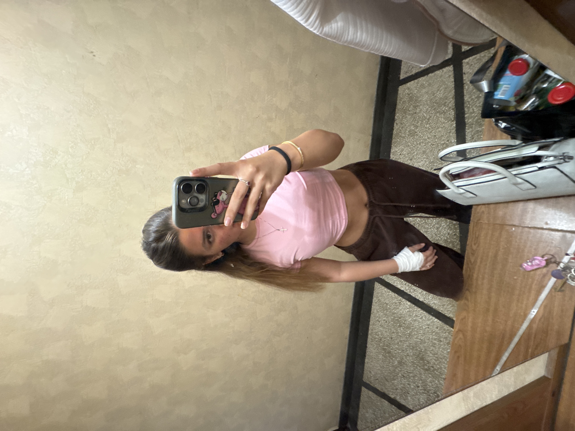 A young woman with long, dark brown hair pulled back is taking a mirror selfie indoors. She is wearing a pink cropped t-shirt and brown sweatpants. Her left hand is wrapped in white medical tape. She is wearing a necklace with a cross pendant, a ring on her right ring finger, and two bracelets on her right wrist. She is holding a smartphone in her right hand, with a pink cartoon character case. The background appears to be a hallway with textured beige walls and a dark tiled floor. A wooden surface is visible in the foreground, with bags and keys resting on it. The location is identified as Sofia, Bulgaria.