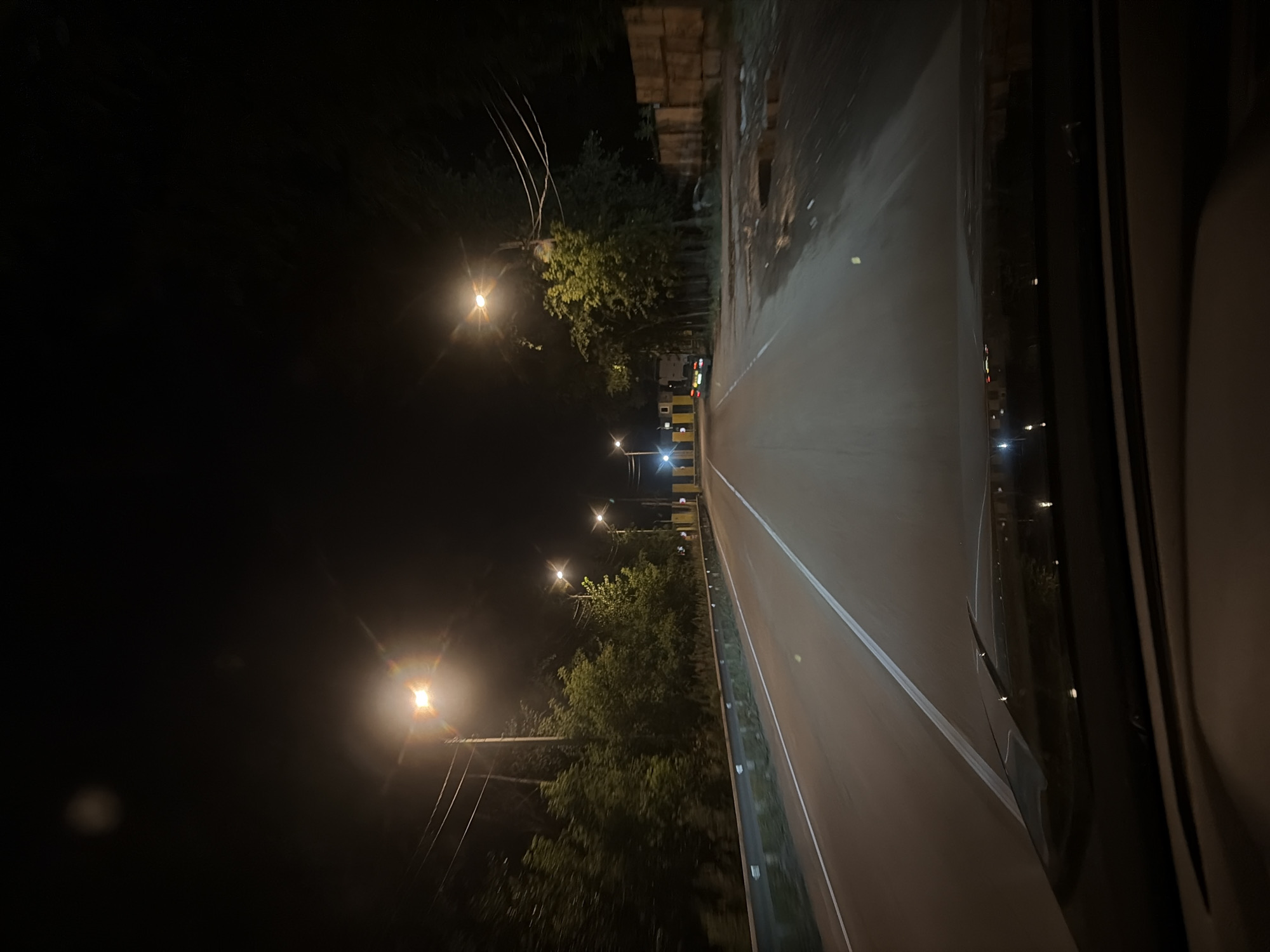 The image depicts a view from a car driving on a two-lane road at night. Streetlights illuminate the asphalt road, which has white dividing lines. On the sides of the road, there are trees and a metal guardrail. In the distance, a yellow structure is visible, possibly a toll booth or a checkpoint. Several vehicles are on the road, and some buildings and wooden structures are on the right side. The sky is dark, indicating nighttime. The location is the city of Elektrougli, Russia.
