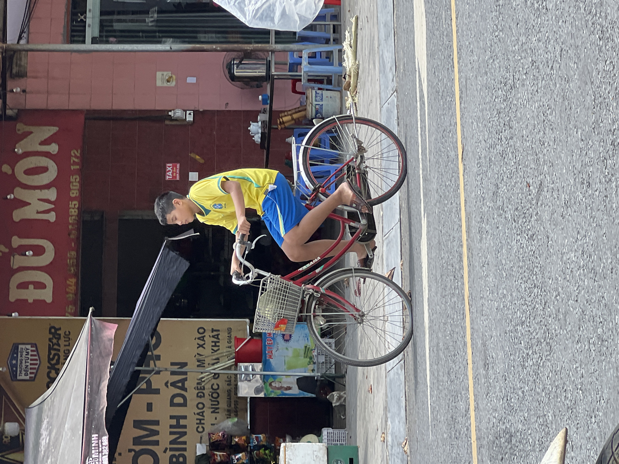 In the city of Tân Quang Commune, Vietnam, a young boy, appearing to be of Vietnamese descent, rides a red bicycle with a basket on the handlebars. He is wearing a yellow t-shirt with a green collar and trim, and blue shorts. His feet are clad in brown flip-flops. The bicycle has a white basket filled with items, including what appears to be a red and orange flower. The scene is set on a paved street with yellow and white markings. In the background, a building with red signage that reads "DÙ MÓN" is visible, along with other storefronts and businesses. There are also blue plastic chairs and tables outside one establishment, and a large metal kettle on a table. The overall environment suggests a bustling street scene in a Vietnamese urban or semi-urban area.