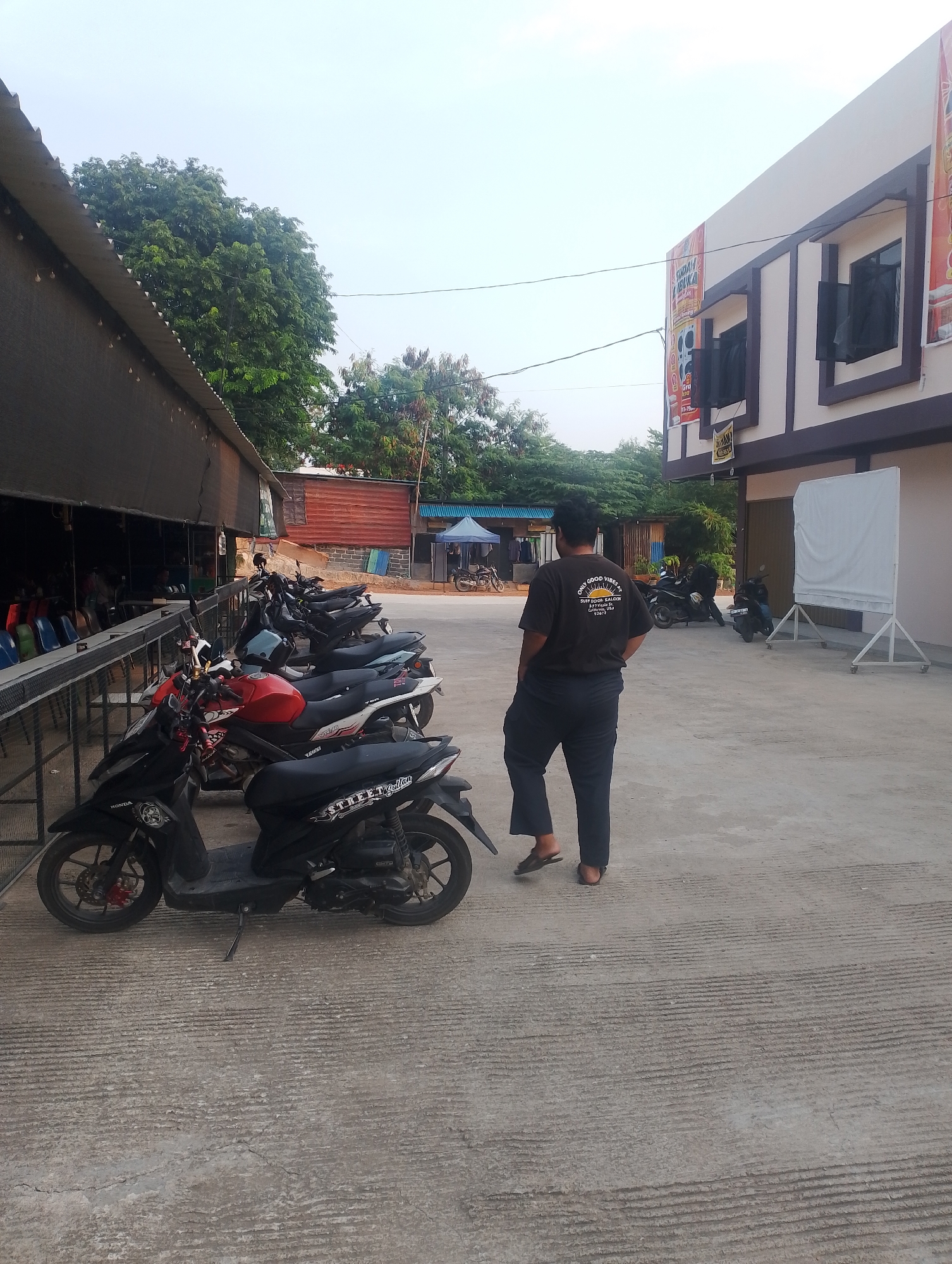 The image depicts an outdoor scene in Batam, Indonesia, likely in front of a building that appears to be a shop or restaurant. A man wearing a dark t-shirt with a sun graphic and text that reads "GOOD VIBES" is standing with his back to the camera, looking towards a row of parked motorcycles. The motorcycles are lined up on a paved surface, with some on a slight incline. To the left of the frame, a long covered structure with seating and tables is visible, suggesting a dining or gathering area. Beyond this structure, trees and smaller buildings are present. To the right, the building facade is visible, featuring windows and signage, including what appears to be a large white screen on a stand. The overall lighting suggests it is daytime, possibly late afternoon.