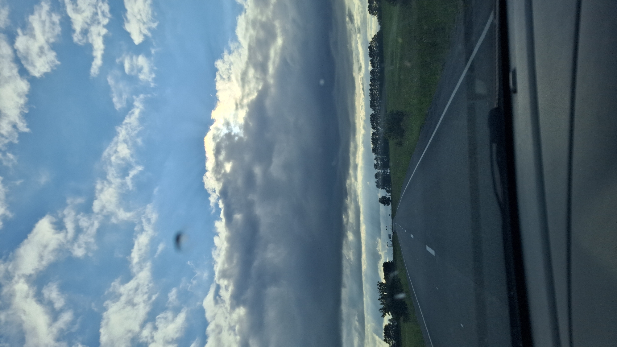 This is a photograph taken from inside a vehicle, looking out the passenger side window. The image is oriented vertically, suggesting it may have been taken from a car in motion or the camera was held at an unusual angle.
The scene is divided into two main parts: the sky on the left and the road and landscape on the right. The sky is a brilliant blue with scattered white clouds, some of which are wispy and others appear more dense and dramatic, tinged with grey. Sunlight is breaking through some of the clouds, creating bright beams. There is a dark, tall cloud formation that dominates the center of the sky, creating a striking contrast. A small, dark, blurry object is visible in the sky, possibly a bug or a speck of dust on the lens.
To the right of the sky, a paved road stretches into the distance, bordered by a strip of green grass and a line of trees. The trees appear to be deciduous and are densely packed, forming a natural barrier along the road. The road itself has white lane markings. The edge of the vehicle, possibly a window frame or door panel, is visible on the far right of the image.
The lighting suggests it is daytime, likely late afternoon or early evening given the dramatic cloud formations and the angle of the sun. The weather appears to be mixed, with patches of clear sky and impending or passing storm clouds. The setting appears to be a rural or semi-rural area, given the presence of the road, fields, and trees. There are no people or explicit text visible in the image. The overall mood is one of movement and a dramatic natural sky.