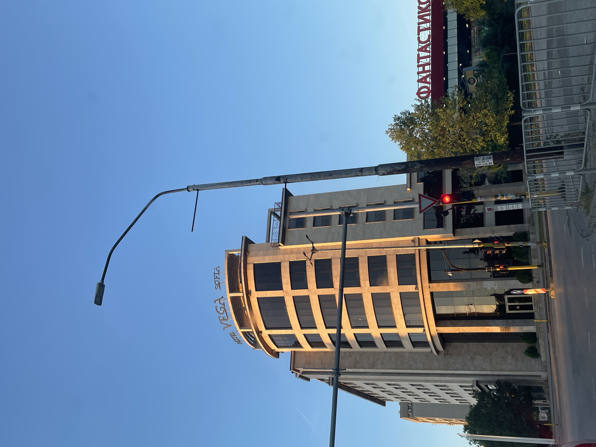 The image depicts a street scene in Sofia, Bulgaria. The main subject is the Hotel Vega Sofia, a modern building with a curved facade and large windows. A street lamp stands in front of the hotel. Across the street, a building with the sign "ФАНТАСТИКО" (Fantastico) is visible. A traffic light is seen displaying a red light. A metal barrier is placed near the curb. The sky is clear and blue, suggesting daylight.