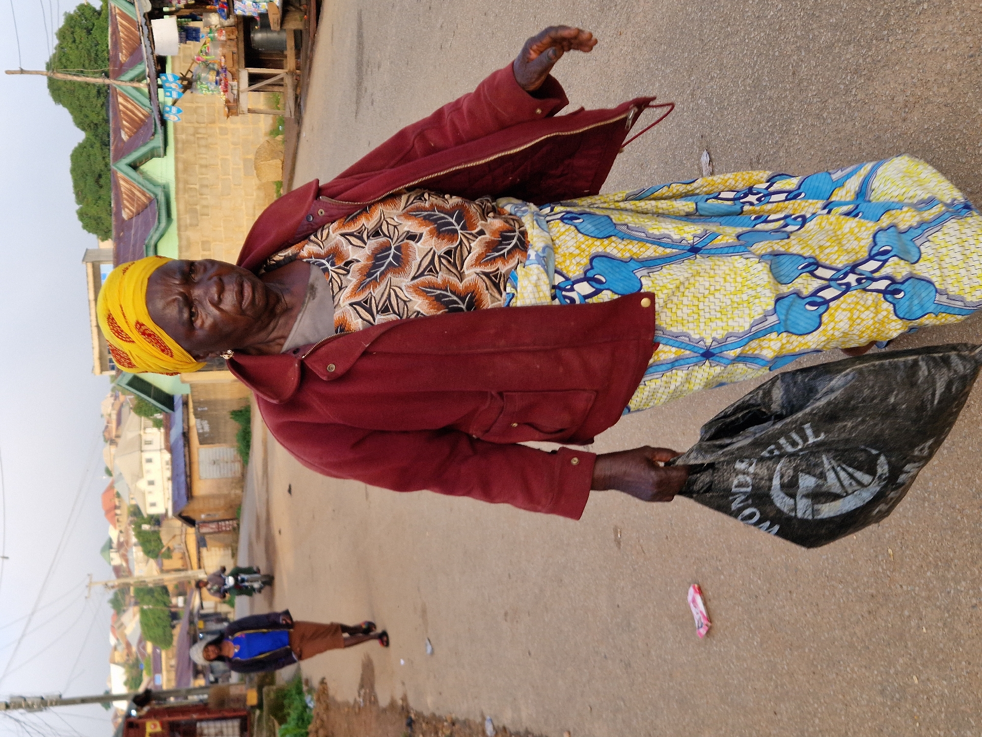 An elderly Black woman with a yellow headwrap walks down a paved road in Kabong, Nigeria. She is wearing a red jacket over a patterned shirt and a brightly colored skirt with blue and yellow designs. She carries a black plastic bag in her right hand. In the background, other people and buildings are visible. A young person in a blue shirt and brown skirt walks away from the camera on the left, and a person on a motorcycle is visible further down the road. Shops and houses line the street, with some structures featuring corrugated metal roofs. The sky is overcast.