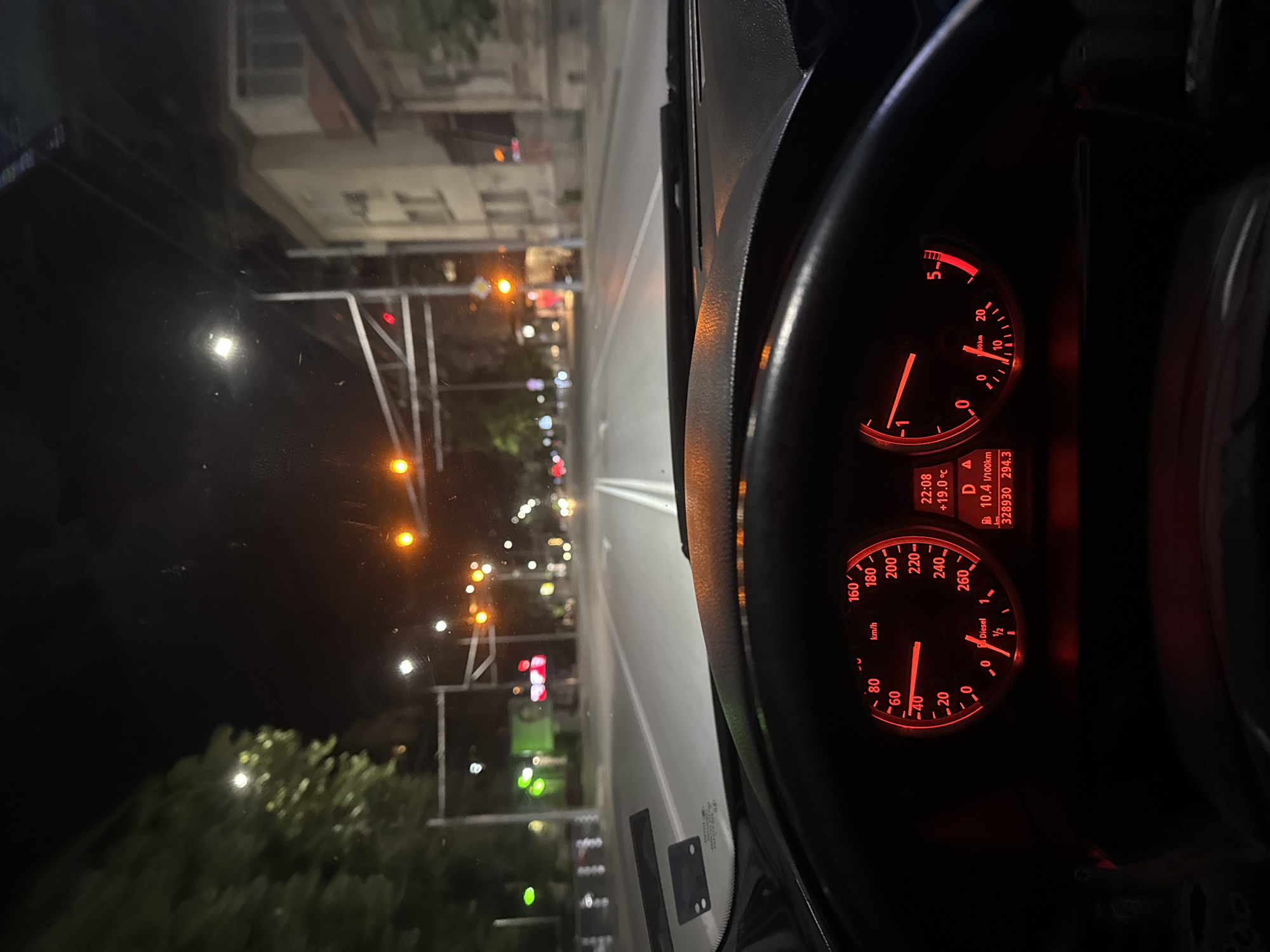 The image depicts a view from inside a car driving on a road at night in what appears to be an urban area. The car's dashboard is visible, with the speedometer and other gauges illuminated in red. The speedometer shows the car is moving at approximately 40 km/h. Outside the car, streetlights illuminate the road and buildings, and some trees can be seen. Based on the streetlights and architecture, the location is likely in Stara Zagora, Bulgaria. The time is 22:08 and the temperature is 19 degrees Celsius.