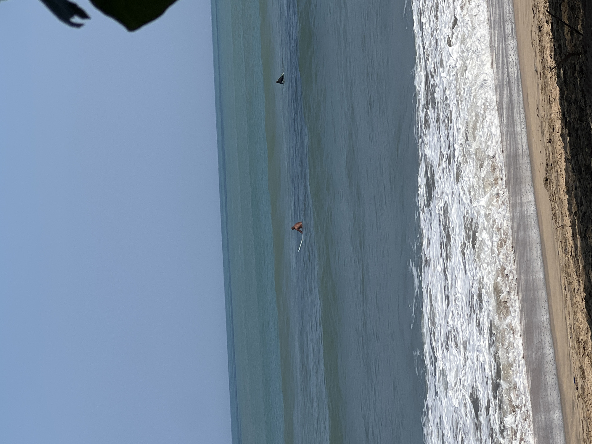 In Kuta, Indonesia, two surfers are visible in the ocean. One surfer, with an orangish-brown complexion, is seated on a white surfboard in the middle of the frame. Another surfer, dressed in dark attire, is further to the right and also seated on a dark surfboard. Both surfers are facing the ocean. The water is a deep blue-green with visible waves and white foam near the shore. The beach in the foreground is sandy with some sparse vegetation. The sky is clear and light blue.