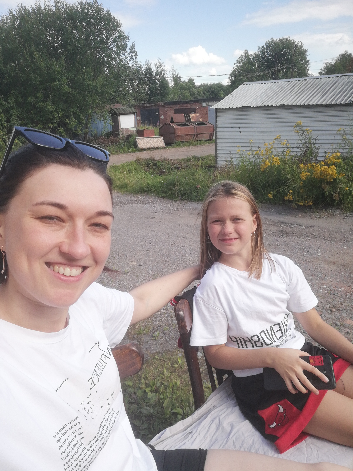 This is a selfie-style photo of two people, likely a mother and daughter, sitting outdoors on a bench. The woman in the foreground is smiling broadly, with sunglasses perched on her head. She is wearing a white t-shirt with text and graphic details. Next to her, a younger girl with blonde hair also looks at the camera, wearing a white t-shirt and black shorts with red trim and a bull logo.
They are in an outdoor setting that appears to be a semi-industrial or rural area, possibly in Tosno, Russia. In the background, there are various structures, including a corrugated metal shed, a brick building, and some rusty industrial containers. Greenery and yellow wildflowers are visible around the bench, suggesting it's daytime and likely summer. The lighting indicates it's a sunny day. The overall mood seems relaxed and happy. The visible text on the women's t-shirts is in a stylized font, partially obscured by the image composition.