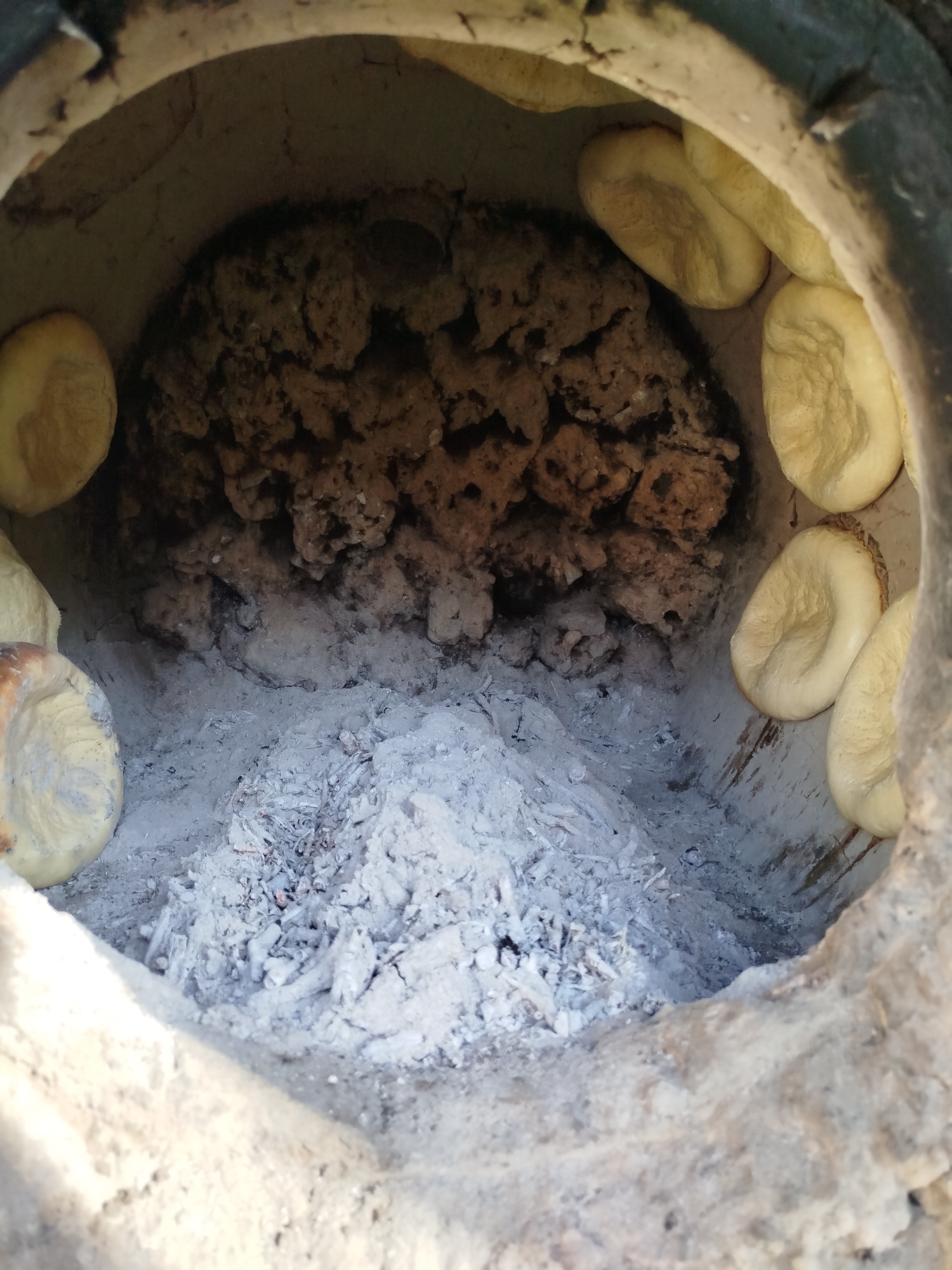The image shows the inside of a tandoor oven, a traditional clay oven used for baking. The tandoor is filled with ash and embers from a fire, with several pieces of flatbread baked onto the inner walls. The bread appears to be light brown and somewhat puffed up. The tandoor itself is made of clay and appears to be well-used. This scene is typical of traditional bread-making practices in rural areas, and this particular tandoor is located in the village of Кызыл-Булак, Kyrgyzstan, which is nestled in the Tien Shan mountains.