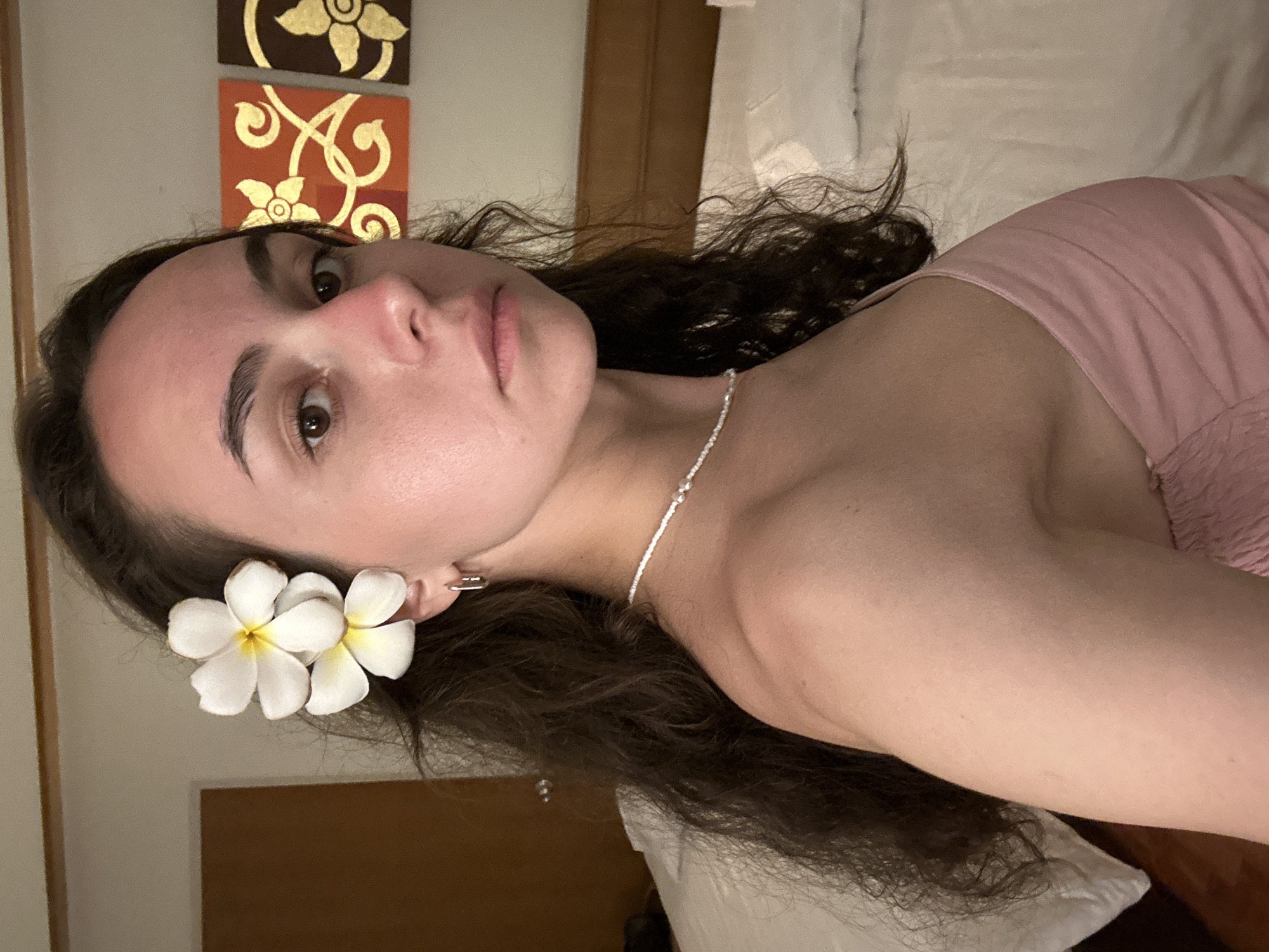 A woman is lying down in an indoor setting. She has dark, curly hair with two white plumeria flowers near her ear. She is wearing a light pink top and a delicate white necklace. The background includes a painting with gold designs and a headboard. The location is Pattaya City, Thailand.