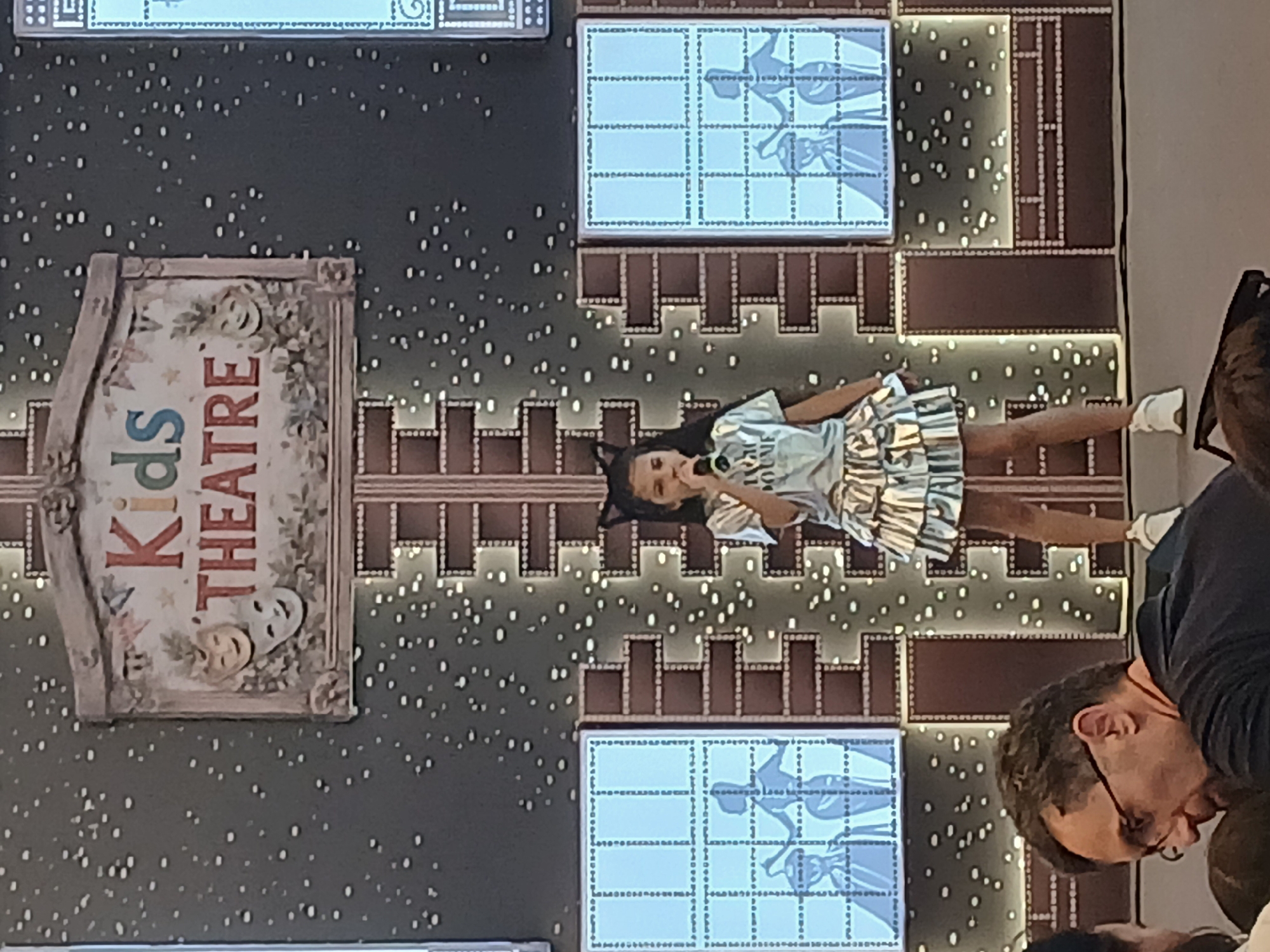 The image shows a young girl in a light-colored, metallic-looking outfit with a ruffled skirt and a matching headband, standing on a stage. She appears to be performing or posing, holding a microphone up to her mouth. Behind her is a dark backdrop speckled with small, bright lights. To the left of the girl is a sign that reads "Kids THEATRE" in colorful lettering, with theatrical masks displayed on it. Above and to the right of the girl, there are large screens displaying what appears to be a black and white image of figures in a grid pattern. The overall setting seems to be a theater or performance space designed for children, with decorative brick-like patterns and glowing lights forming the backdrop. The lighting is bright on the stage, illuminating the girl and the immediate surroundings. The image is oriented sideways.