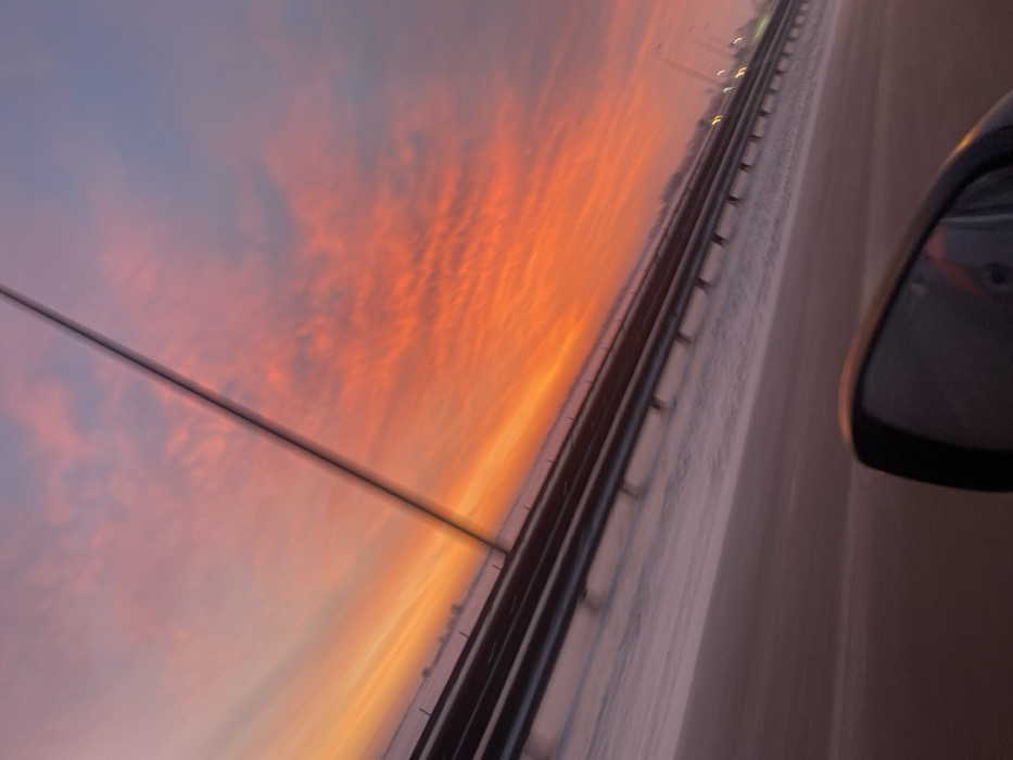 From a car window perspective, a vibrant sunset sky transitions from pale lavender to fiery orange and red, streaked with wispy clouds. The view is framed by the vehicle's side mirror and a blurred highway barrier. A power line stretches across the upper left quadrant. The road, barely visible on the right, is a light, neutral color. The shot conveys a sense of motion and transient beauty.