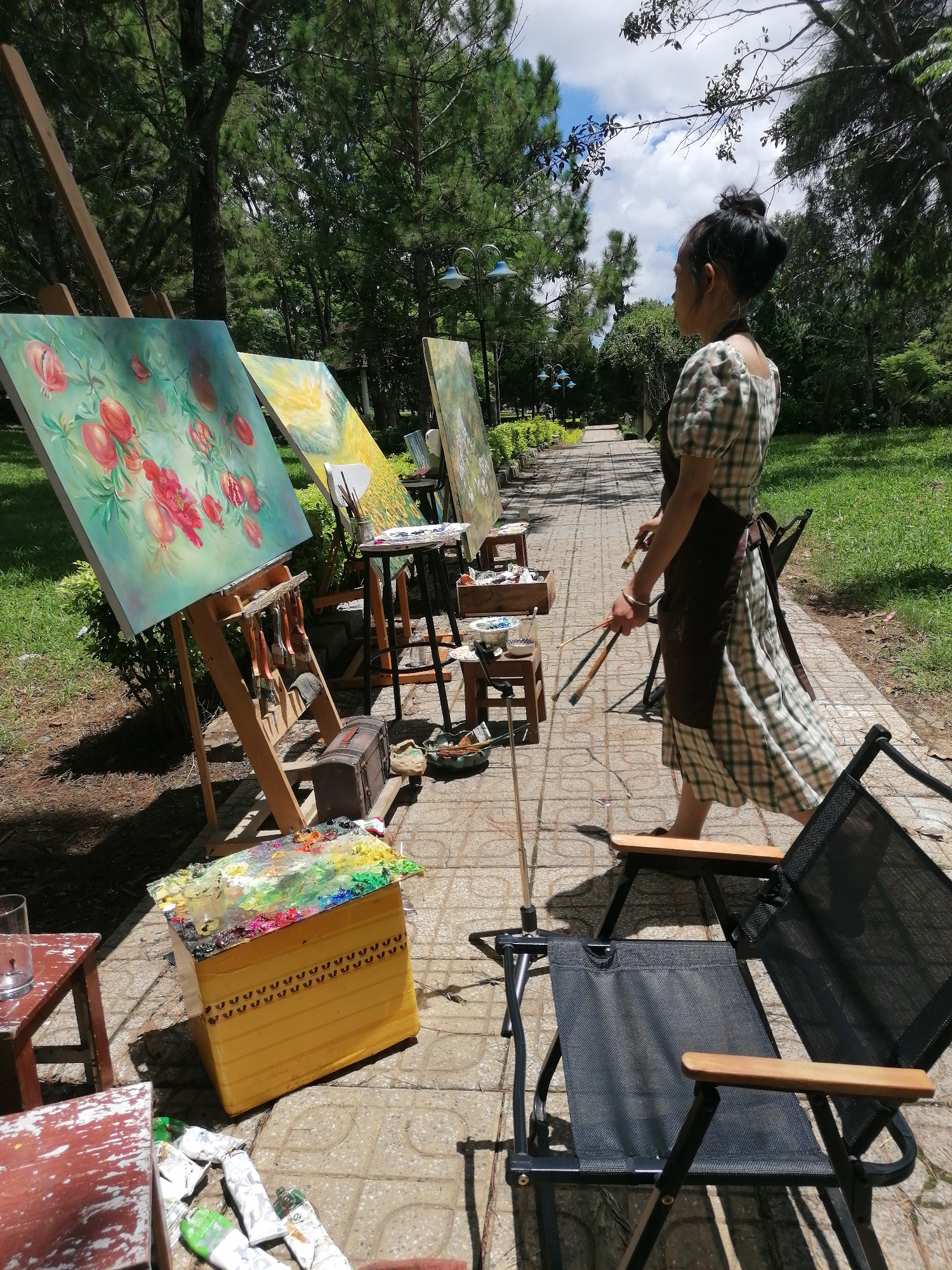 An outdoor painting setup is arranged along a paved walkway in Da Lat, Vietnam. The scene features several easels displaying oil paintings, predominantly still lifes of fruits and flowers. A woman, dressed in a plaid dress and apron, stands with paintbrushes in hand, facing away from the camera and towards the paintings. Surrounding the easels are various art supplies, including paint palettes, tubes of paint, brushes, and small wooden stools. Two folding chairs are positioned on the walkway, one occupied by the woman and another empty. The environment is characterized by lush greenery, tall pine trees, and a bright, cloudy sky, suggesting a park or garden setting.