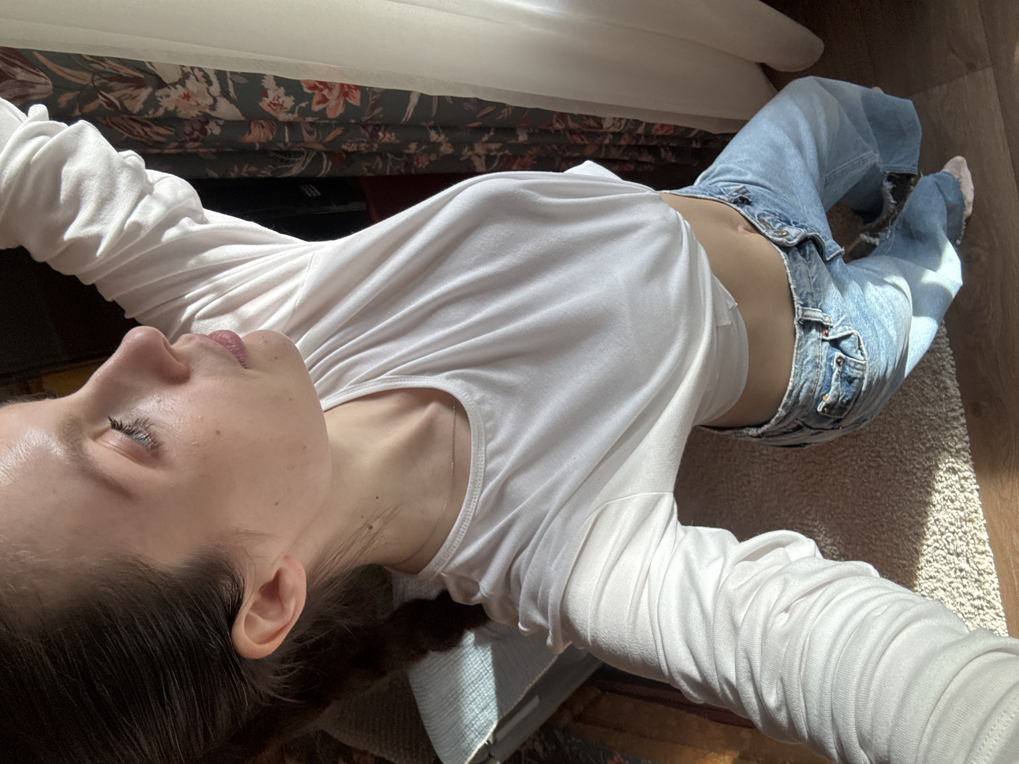 A woman lies on a carpeted floor indoors, presumably in Tomilino, Russia. She wears a white long-sleeved shirt and light-wash jeans. Her arms are outstretched. Her hair is dark and her eyes are closed. Curtains are visible in the background. The lighting suggests it is daytime.