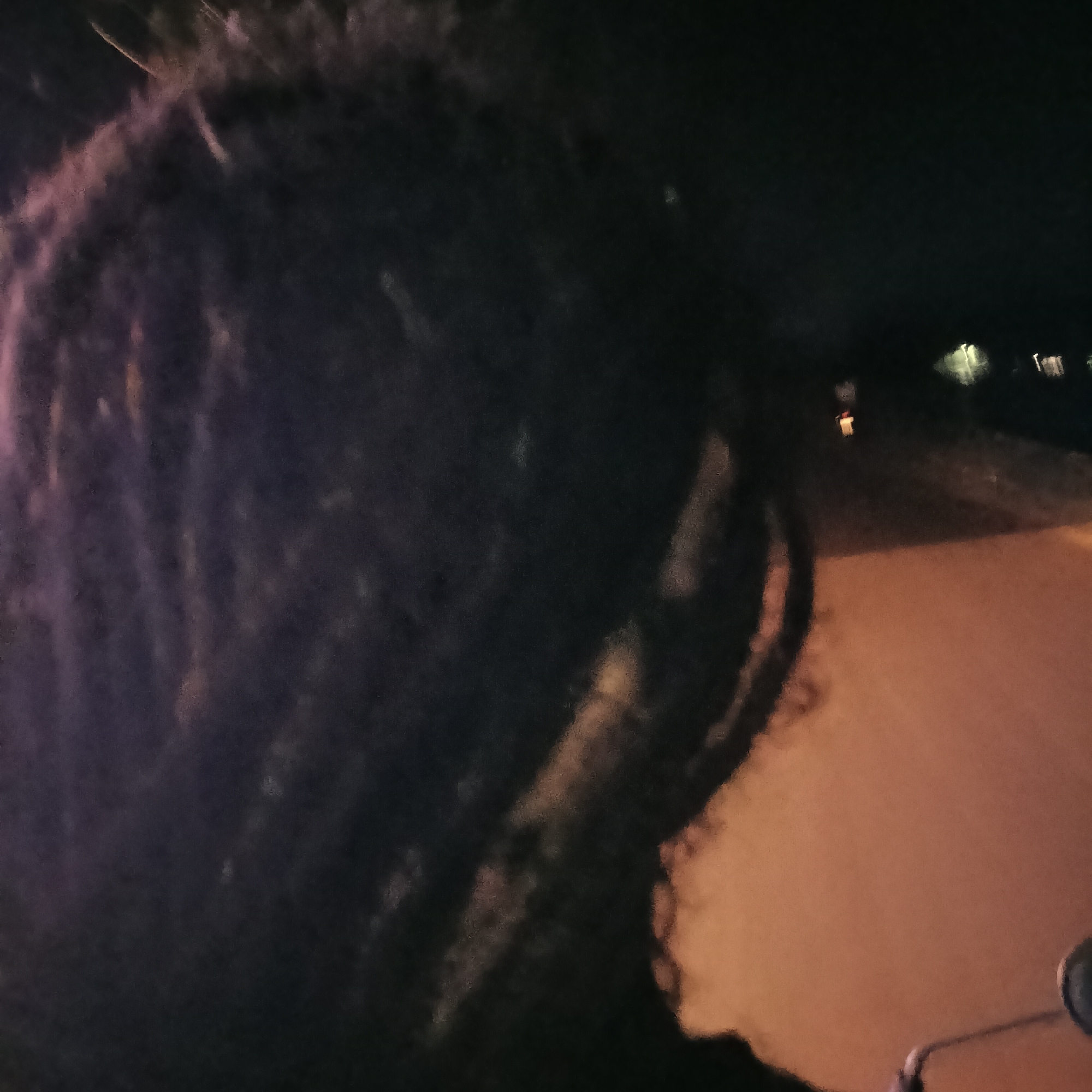A blurry, low-light photograph appears to depict a person with dark dreadlocks, possibly on a motorcycle. The background includes a dimly lit road with streetlights and a suggestion of roadside structures. The location is unknown but, based on the user query, it is purported to be in Oxel, India. Due to the image quality, details are difficult to ascertain.