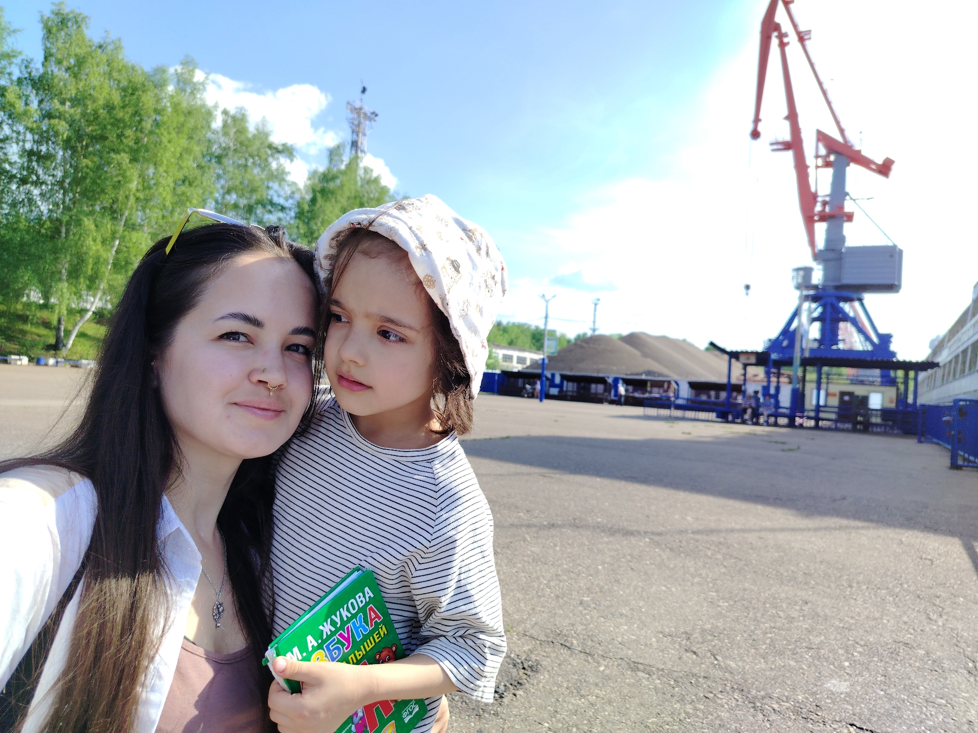 The image is a medium shot of a woman and a young girl, appearing to be a mother and daughter, taking a selfie in an urban industrial setting. The woman has dark hair, a nose ring, and is wearing a light-colored blouse over a darker top. She's holding the phone and looking at the camera with a slight smile. The young girl, wearing a striped shirt and a patterned bonnet, is looking off to the side. She's holding a green book with Russian text, "М.А. Жукова Букварь для малышей."
In the background, there's a port crane with red and blue elements, indicating a harbor or shipping area. There's also a pile of material, possibly coal or gravel, and a building with blue metal fencing and canopies. The sky is blue with some clouds, suggesting a bright, sunny day.
Based on the book and local architectural details, the location is likely in the city of Chaikovsky, Russia, considering the book title and the types of industrial facilities often found near Russian river ports.