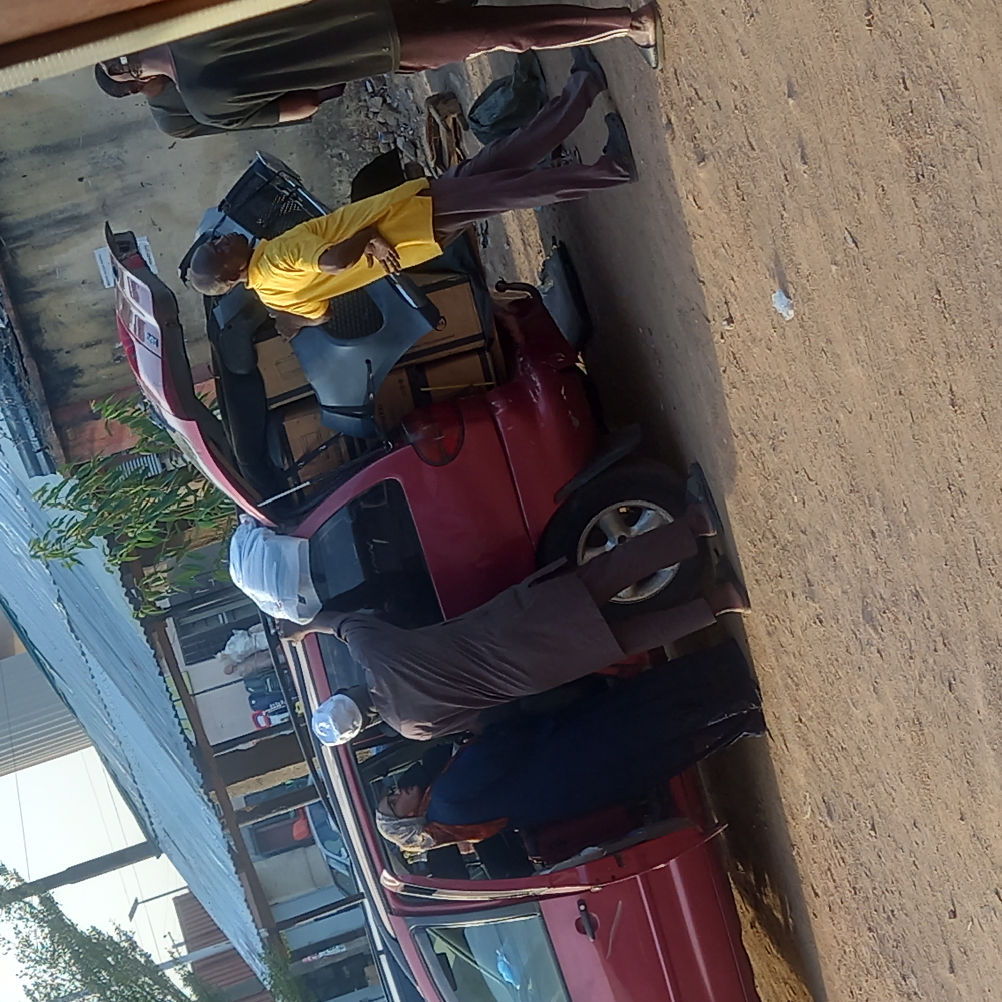The image, taken in Dadi Riba, Nigeria, captures an outdoor scene with several men working around a small, red two-door car on a sunny day. The ground is unpaved, consisting of dirt or sand, and natural light suggests it's daytime with clear weather.
At least five men are visible. In the upper left, a man in a dark shirt and cap stands, observing the activity. A man in a bright yellow t-shirt and a light cap is positioned near the rear of the red car, which has its trunk or hatchback open. He appears to be handling black car parts, possibly a bumper or fender, along with cardboard boxes being loaded or unloaded.
Another man, wearing a dark brown/grey long-sleeved shirt and a white cap, is leaning into the open driver's side door of the car. Inside the passenger compartment, a person in a blue jacket/shirt and a white cap/hairnet is visible. The legs of a fifth individual in dark pants and shoes are seen near the rear wheel on the passenger side.
The red car is a compact model, and the activity centers on its interior and rear, suggesting either loading/unloading goods or vehicle maintenance. In the background, buildings with corrugated metal roofs and some green foliage are visible. No legible text is discernible on the car or other objects. The scene depicts a busy, practical activity in an open-air setting.