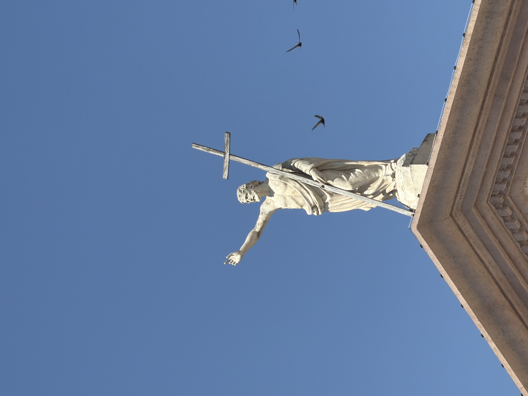 A stone statue of Jesus Christ stands on the roof of a building in Peschiera del Garda, Italy, against a clear blue sky. Jesus is depicted with a beard and is wearing a robe, holding a wooden cross in his left hand and pointing with his right. Three birds are in flight in the background, to the right of the statue. The building's roof is made of stone with decorative elements.