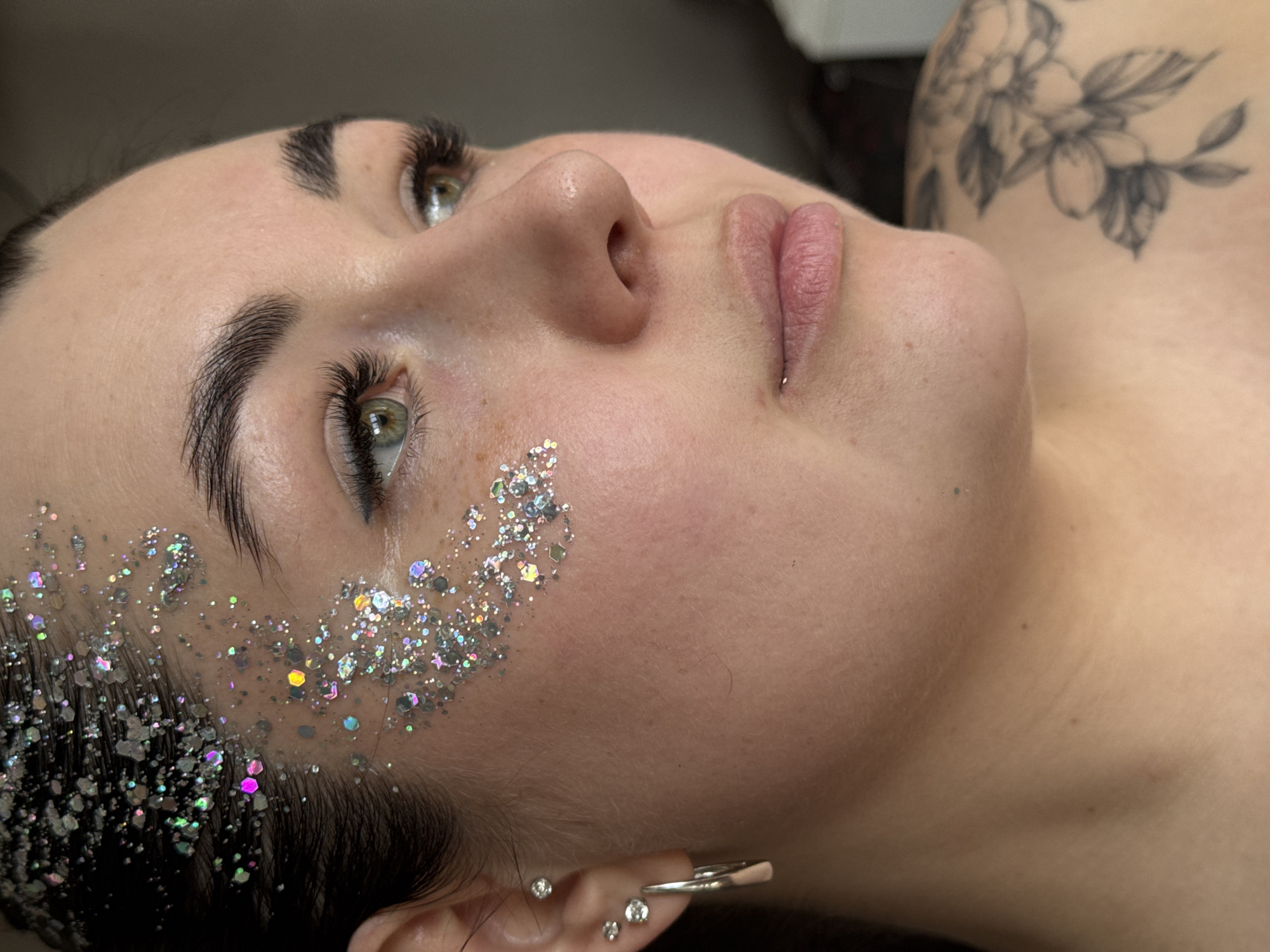 The image presents a close-up portrait of a person, likely female, positioned with her head tilted back, looking upwards and to the right of the frame. Her face, seen from a low-angle profile, is the primary subject. She possesses fair, smooth skin, bright green or hazel eyes with prominent, dark eyelashes, and well-defined, dark eyebrows. Her lips are slightly parted, showcasing a natural pink color.
A notable feature is the artistic application of chunky, iridescent silver glitter on her left temple and cheekbone, extending into her dark hair. The glitter reflects various hues, including pink, blue, yellow, and green, suggesting a holographic effect. Her right ear is partially visible, adorned with multiple small, clear stud earrings in the lobe and helix, and a silver hoop or cuff higher up.
A black and white floral tattoo, featuring flowers and leaves, is visible on her left shoulder or upper arm. The background is indistinct, appearing as a plain, light surface, indicating an indoor setting. The media appears to be a posed beauty shot, possibly showcasing makeup artistry, eyelash extensions, or a festive look. There is no visible text. The context for this media is Chaikovsky, Russia.
