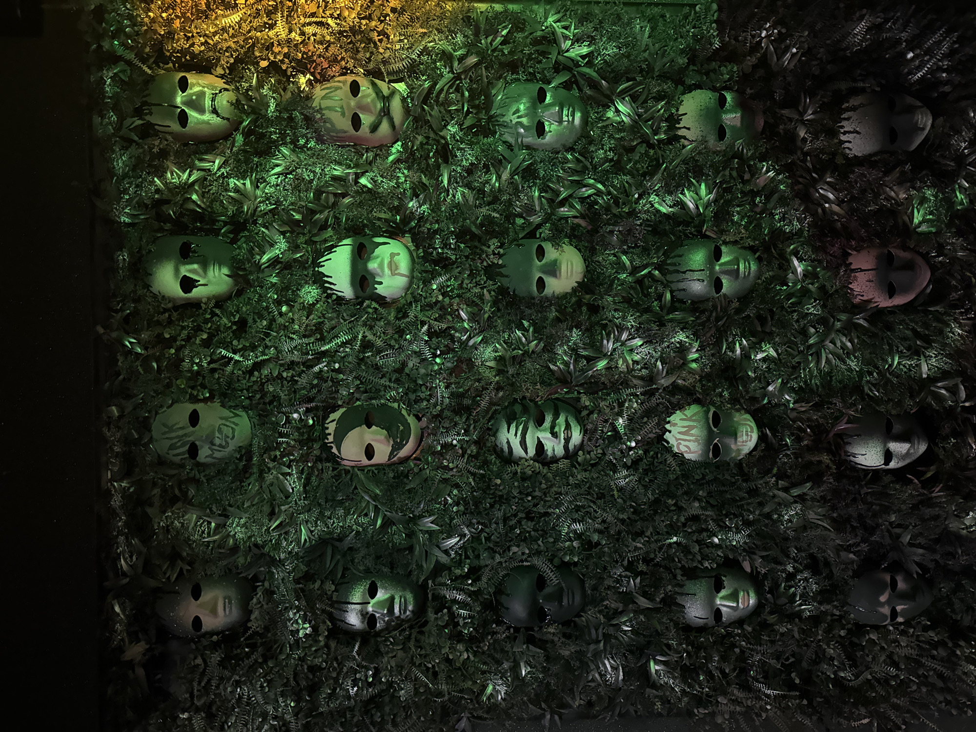 The image shows a wall covered in artificial greenery and decorated with a grid of masks. The masks are face-shaped and appear to be made of a light-colored material with varying designs painted on them. The lighting is uneven, with the top left corner being brightly illuminated, casting a yellowish hue, while the right side is darker. The background consists entirely of artificial foliage, creating a dense, textured appearance. The scene is likely an artistic installation or decorative feature, possibly located in Plovdiv, Bulgaria.