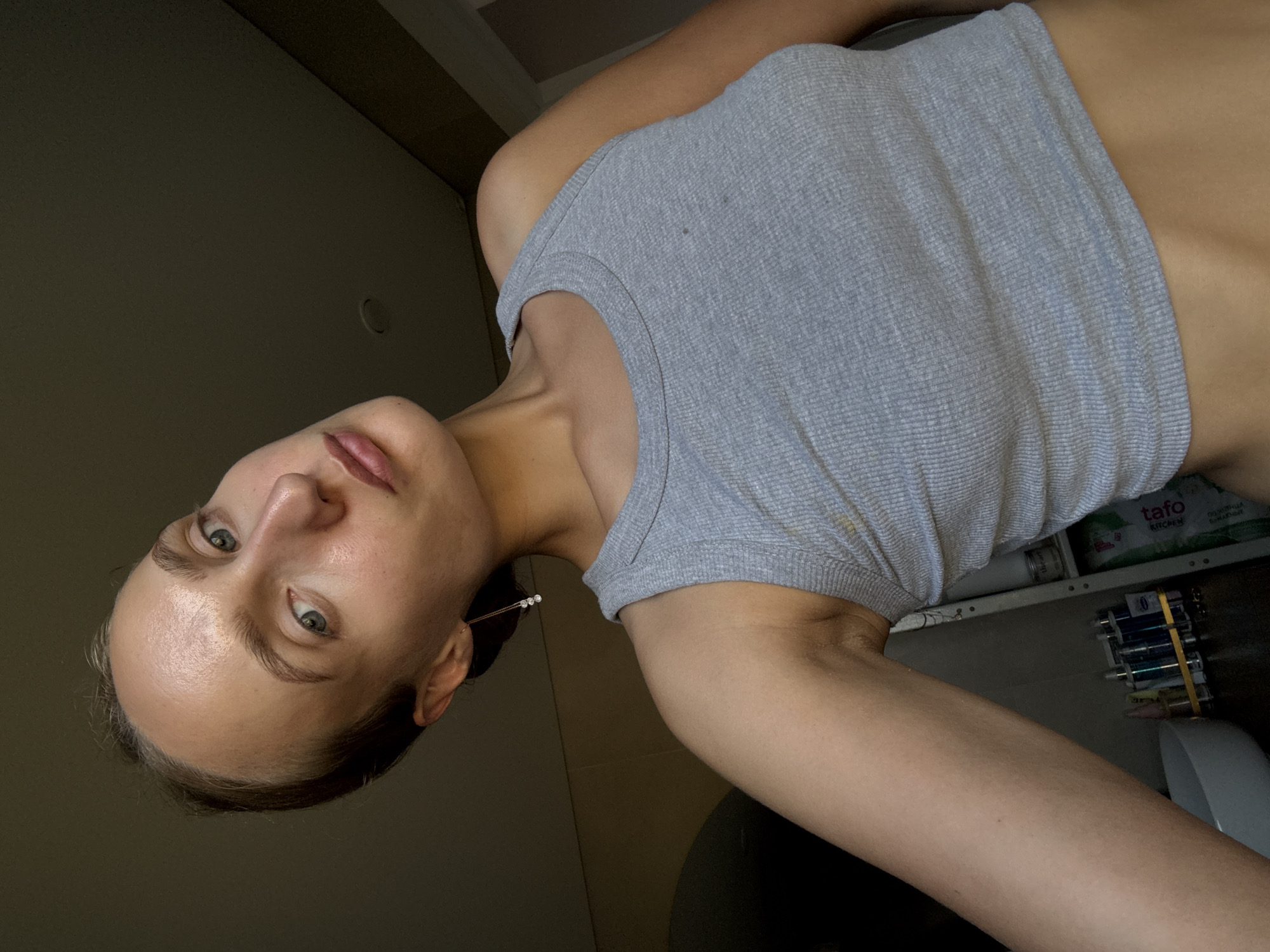 A young Caucasian woman with light skin and blonde hair is taking a selfie in what appears to be a bathroom. She is wearing a grey ribbed tank top. Her head is tilted to the side, and she is looking towards the camera with a neutral expression. Her left arm is extended, holding the phone. Her right shoulder and part of her chest are visible. The background is dimly lit and shows a wall with some shelves and bottles, suggesting a bathroom or vanity area. The lighting suggests artificial light, likely from above or to the side, creating some highlights on her face. There is no visible indication of the city of Подшивалово, Russia.