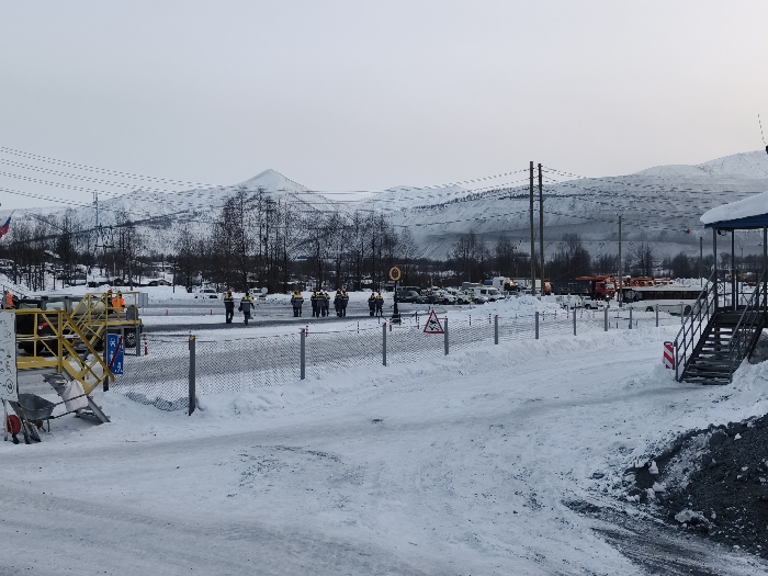 The image displays a snow-covered industrial or mining complex in the city of Im. Gastello, Russia, under an overcast, light gray sky.
In the foreground, an extensive snow layer covers the ground, showing tire tracks on a cleared path that extends into the midground. To the far left, a yellow metal industrial platform or scaffolding structure is present, with two individuals in orange high-visibility jackets and hard hats visible near it. A wheelbarrow rests below the platform. A partially visible flag with white, blue, and red horizontal stripes is positioned behind this structure.
A wire mesh fence, supported by metal posts, stretches horizontally across the mid-left foreground, separating the immediate area from a paved lot. Attached to this fence is a blue sign with white pictograms, possibly indicating a facility. Beyond the fence, a group of approximately eight to ten individuals, all uniformly dressed in high-visibility vests (yellow or orange) and hard hats, walk in a line across a partially cleared, snow-dusted paved surface, moving from left to right across the frame.
The midground features a large parking or staging area, also partially covered in snow. Numerous vehicles are parked here, including multiple white vans, various trucks, a prominent red fire truck, and a white passenger bus. Several tall utility poles with multiple overhead power lines span horizontally across the midground. Sparse, bare deciduous trees are visible between the industrial structures and the distant mountains.
On the right side of the foreground, a snow-covered mound of dark, granular material is present. Adjacent to it, a dark-colored wooden staircase with metal handrails ascends to a small, elevated structure with a blue roof and open sides. Various warning signs are visible throughout the scene, including a triangular sign with a red border and black pictogram (suggesting caution) and a circular sign with a red border.
The background is dominated by a range of large, snow-covered mountains and hills, stretching across the horizon under the uniformly gray sky, with a single prominent peak visible towards the upper-center-left.