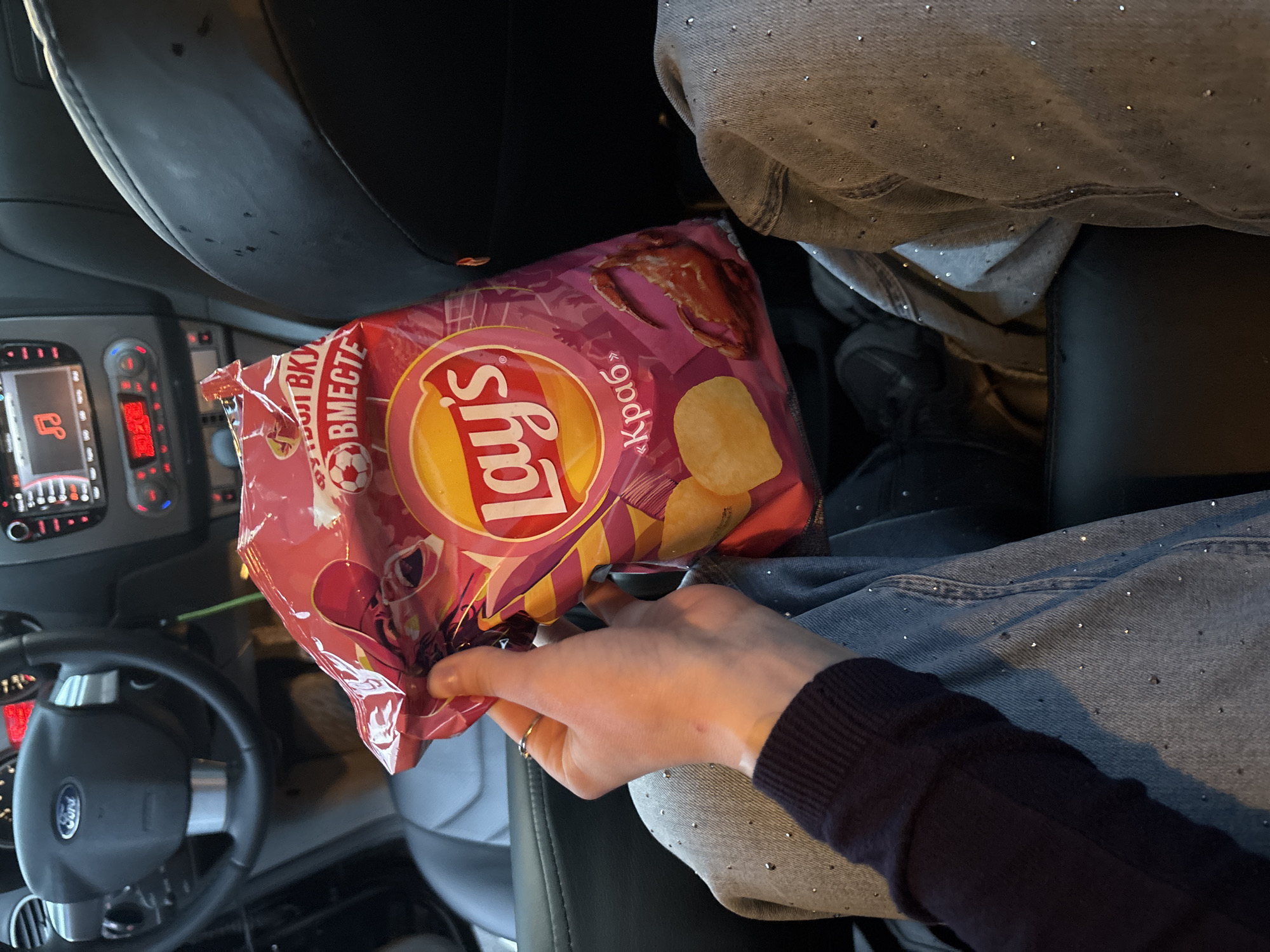 An individual is holding an open bag of Lay's crab-flavored potato chips in the driver's seat of a car. The steering wheel and dashboard of the car are visible in the background. The individual is wearing a dark blue long-sleeved shirt and light blue jeans with rhinestones. The car appears to be parked, and the overall lighting suggests it is daytime. The scene is indoors, within a vehicle, and no specific landmarks of the city of Arfa, Russia are depicted.