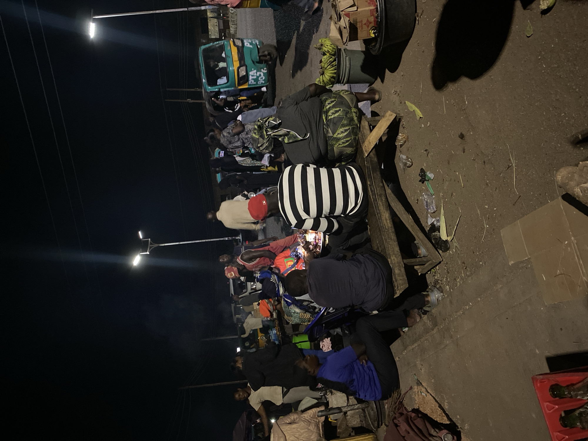 In Bukuru, Nigeria at night, a group of people gather around a low wooden structure, possibly a makeshift table or bench, under streetlights. Individuals are either sitting or standing close to each other, some appearing to be engaged in an activity that involves small objects, potentially gambling or a similar game of chance. A green and yellow auto rickshaw (tuk-tuk) is parked nearby. A bucket containing green bananas rests beside the seated people. Debris and litter scatter the ground.