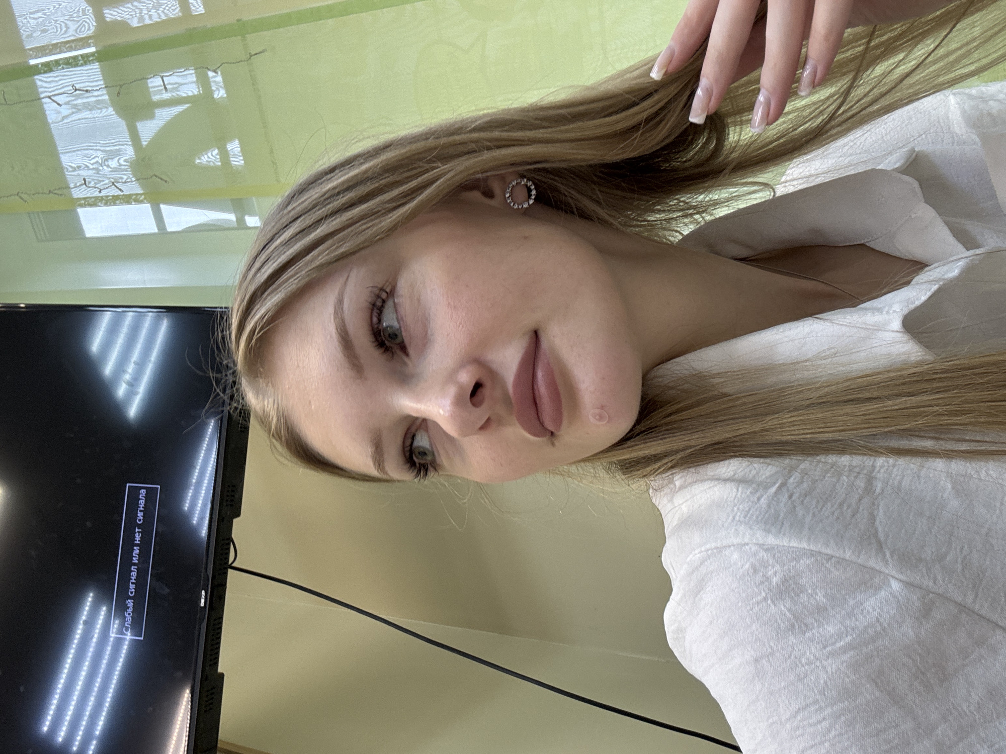 A young woman with blonde hair and light makeup is shown in a close-up shot. She is wearing a white collared shirt. In the background, a dark television screen displays a message in Russian: "Слабый сигнал или нет сигнала" which translates to "Weak signal or no signal." A black cable is visible running vertically on the left side of the screen. A green curtain or drape is on the right side of the image, with a window partially visible behind it. The lighting suggests an indoor setting, possibly an office or a room in an apartment. The overall impression is that the woman is either in her own space or a professional environment where the television is not functioning correctly. There is no indication of the specific location within the city of Perm, Russia.