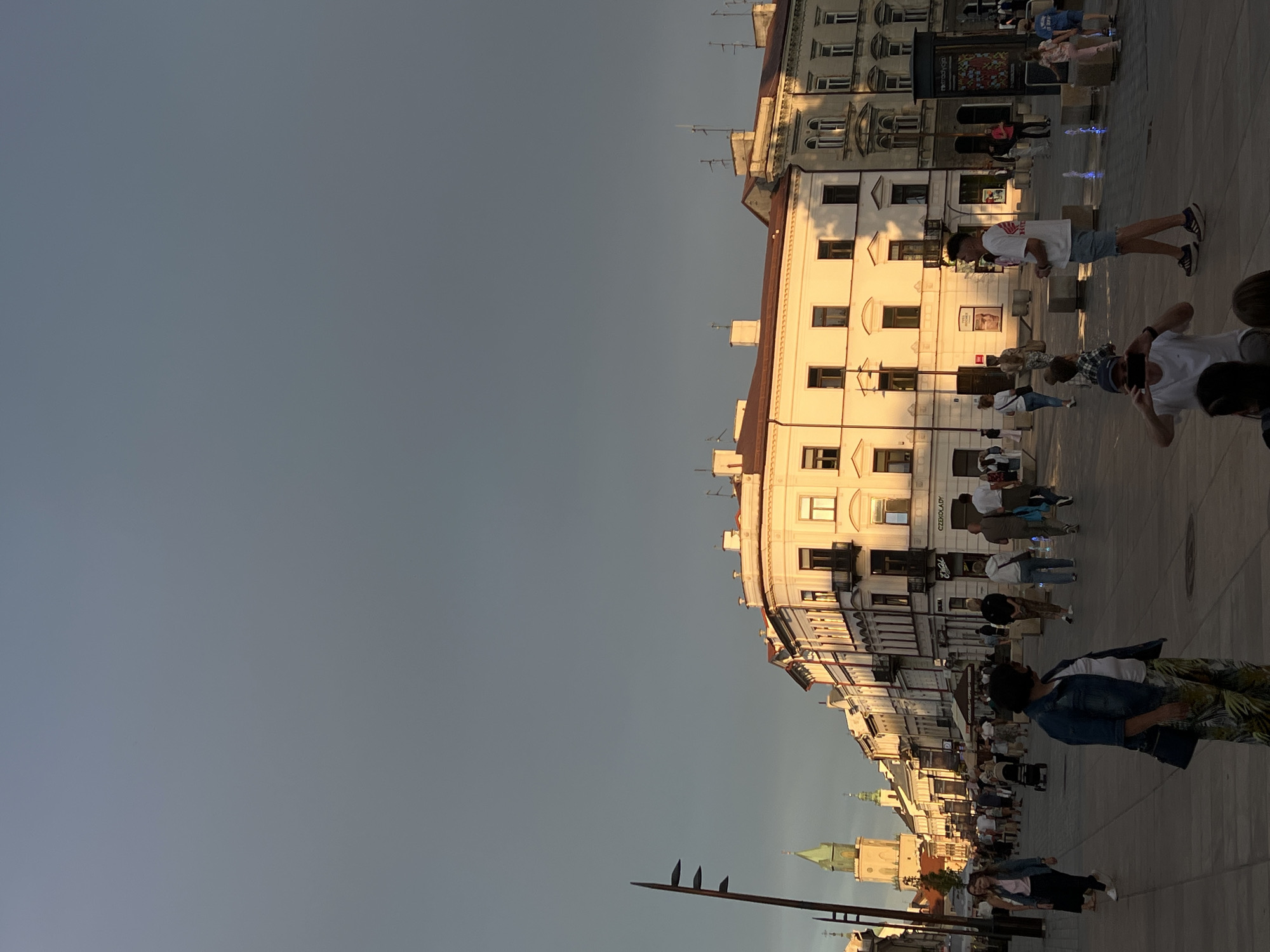 The image depicts a street scene in Lublin, Poland, during what appears to be late afternoon or early evening, indicated by the warm, low-angle sunlight casting long shadows. The architectural style of the buildings suggests a historic European city center.
In the foreground, several individuals are visible. A woman in a denim jacket and patterned pants is standing and looking to her right. To her left, a woman in a black skirt and pink top is walking away from the camera. Further back, a man in a white t-shirt and denim shorts is facing left, speaking into a microphone. In the lower left corner, a person is holding a smartphone and taking a photo or video. Children are also present in the mid-ground, with one child in pink pajamas and another in blue shorts.
The street is paved with stone tiles, and several planters and small fountains are distributed across the area. Buildings line both sides of the street, featuring classical architecture with multiple stories, arched windows, and balconies. One prominent building in the background has a green-spired tower. The sky is a gradient of dark gray at the top, transitioning to lighter hues near the horizon where the sun is illuminating the buildings.
The atmosphere appears to be relaxed, with people strolling and engaging in various activities, suggesting a public square or pedestrianized street. The presence of shops with signage, such as "CHOCOLAT," further indicates a commercial area.