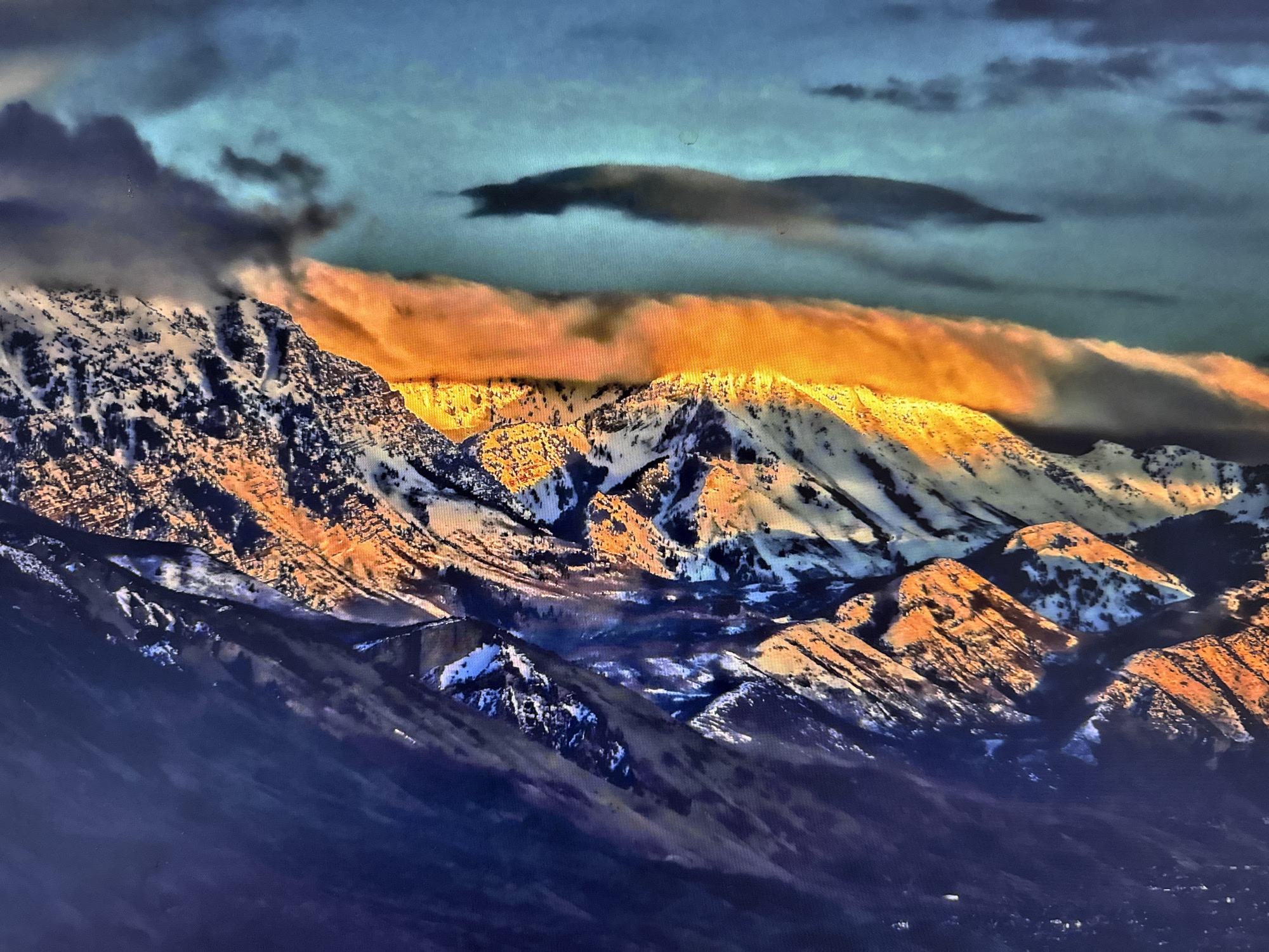 The image presents a dramatic mountainous landscape, likely at sunrise or sunset, showcasing a striking contrast between warm and cool tones. A vast range of snow-capped peaks dominates the scene, their upper sections and crests brilliantly illuminated by a golden-orange light. This warm glow highlights the rugged textures of the rock and snow, casting deep blue-purple shadows into the valleys and unlit slopes below.
Above the mountain range, the sky is a dynamic canvas of clouds. A prominent, elongated band of clouds, mirroring the golden-orange light on the mountains, stretches horizontally across the mid-sky, seemingly hovering just above the peaks. Above this luminous layer, the sky transitions to a cooler blue-green hue, with darker, more turbulent clouds scattered throughout, including a distinct, dark, horizontal cloud formation in the upper sky.
The scene conveys a sense of serene grandeur and the raw power of nature. The presence of snow indicates cold weather conditions, typical of a mountain environment. There are no people visible, and no specific activity is taking place beyond the natural phenomena of light and weather. No discernible text is present in the image. This majestic mountain view is situated in the region of Tobolsk, Russia.