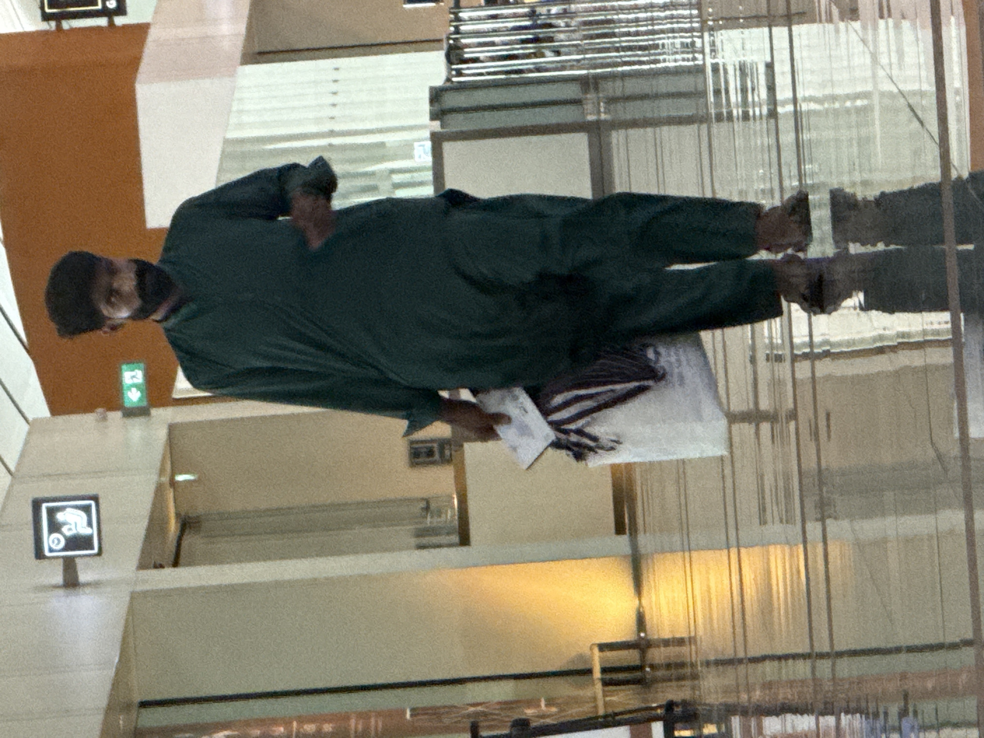 A man is walking through an indoor space with polished floors that reflect the light and surroundings. He is wearing a dark green traditional outfit consisting of a long tunic and loose trousers. In his right hand, he is carrying a white bag with striped fabric visible inside, and a piece of paper. He appears to be walking towards the viewer. The background includes a wall with signage, and vertical architectural elements. This scene is located in the Muscat International Airport in Seeb Province, Oman.