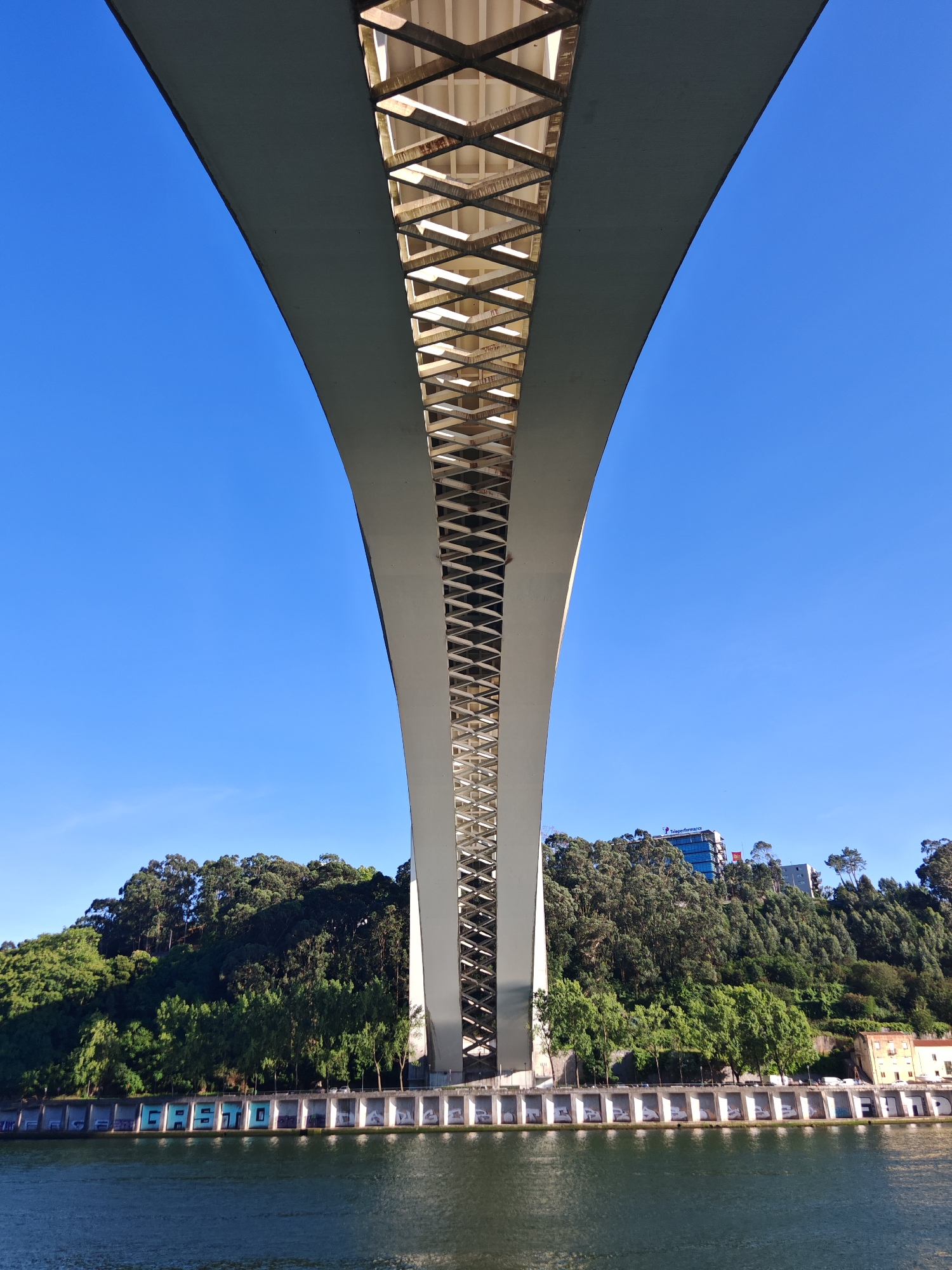 The image captures a low-angle view looking up at a large, modern bridge spanning across a body of water. The underside of the bridge is visible, revealing a complex lattice of white metal beams and concrete structures. The bridge arches upwards, disappearing into a clear, bright blue sky.
Below the bridge, a tree-covered hillside rises steeply. The trees are lush and green, with some buildings visible at the top of the hill, including a modern glass-fronted building with the text "TripeRformance" and a red accent. Along the water's edge, at the base of the hill and beneath the bridge, is a row of white, arched structures, some of which are decorated with graffiti. The water in the foreground is dark and reflects the structures and the sky. The overall scene suggests an urban environment, likely a riverfront in Porto, Portugal, given the architectural style of the bridge and the surrounding greenery. There are no people or animals clearly visible in the image.