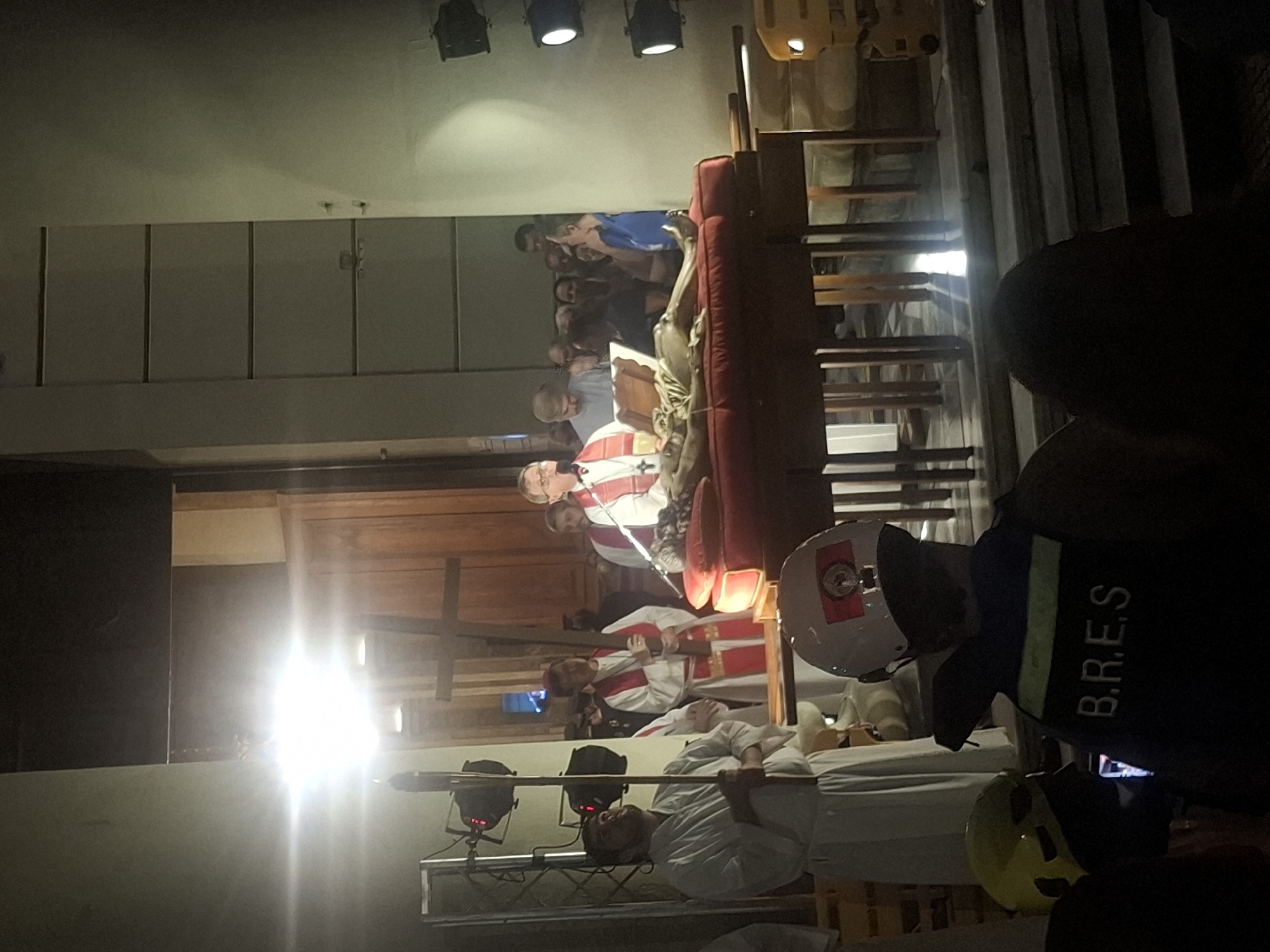 The image depicts a scene inside a building, likely a church or cathedral. Several men in religious attire are present, including at least three wearing red and white robes, possibly cardinals or bishops. One man is lying on a stretcher-like bed. Firefighters in helmets and vests are also present, with one vest visibly labeled "B.R.E.S." A crowd of people is visible in the background. The location is likely Buenos Aires, Argentina, given the Spanish initials on the firefighter's vest and possible relatedness to local events. There is equipment with lights. The scene may depict a religious figure receiving medical attention, or perhaps a rehearsal of a religious ceremony.