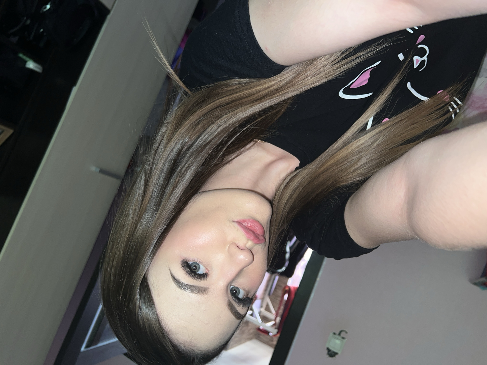 A young woman with long, straight, brown hair is looking directly at the camera. She has light skin, blue eyes, and pink lips. She is wearing a black t-shirt with a white cat design with pink details. The background shows a room with a black cabinet on the left and a glimpse of another room with a chair and a red element. The environment appears to be indoors. The location, Noviy, Russia is inferred and cannot be visually confirmed.