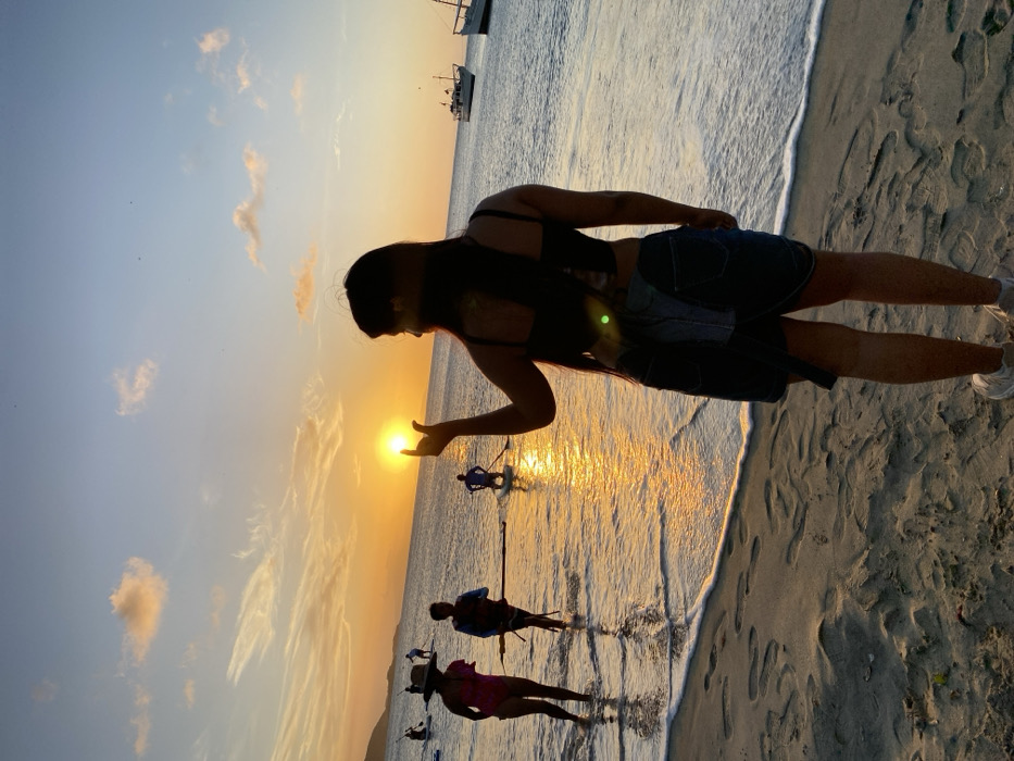 The image depicts a sunset scene on a sandy beach located in Juan Griego, Venezuela.
In the immediate foreground, a female subject is viewed from behind, standing on the wet sand. She has long, dark hair and is dressed in a dark, possibly black, spaghetti-strap top, a denim skirt or shorts, and light-colored sneakers. Her right hand is raised and positioned to visually "hold" the disc of the setting sun, which is a bright orange-yellow sphere directly on the horizon line.
The sky transitions from a light blue at the top to vibrant orange and yellow hues near the horizon, with scattered, thin clouds present. The calm ocean water in the midground and background reflects these sunset colors, and gentle waves are observed breaking on the shoreline.
In the midground, several individuals are present in the shallow water. To the left, a person wearing a wide-brimmed hat and a pink swimsuit is wading. Adjacent to them, another individual, potentially wearing a life vest, is also wading and holding a long paddle or pole. Further out in the water, at least four other individuals are discernible as silhouettes on stand-up paddleboards, with some actively paddling.
In the far background on the right side of the image, two small to medium-sized boats, appearing white with dark hulls, are anchored or floating in the water. Distant landforms, visible as dark silhouettes of hills or mountains, frame the left side of the horizon. The sandy beach shows visible footprints.