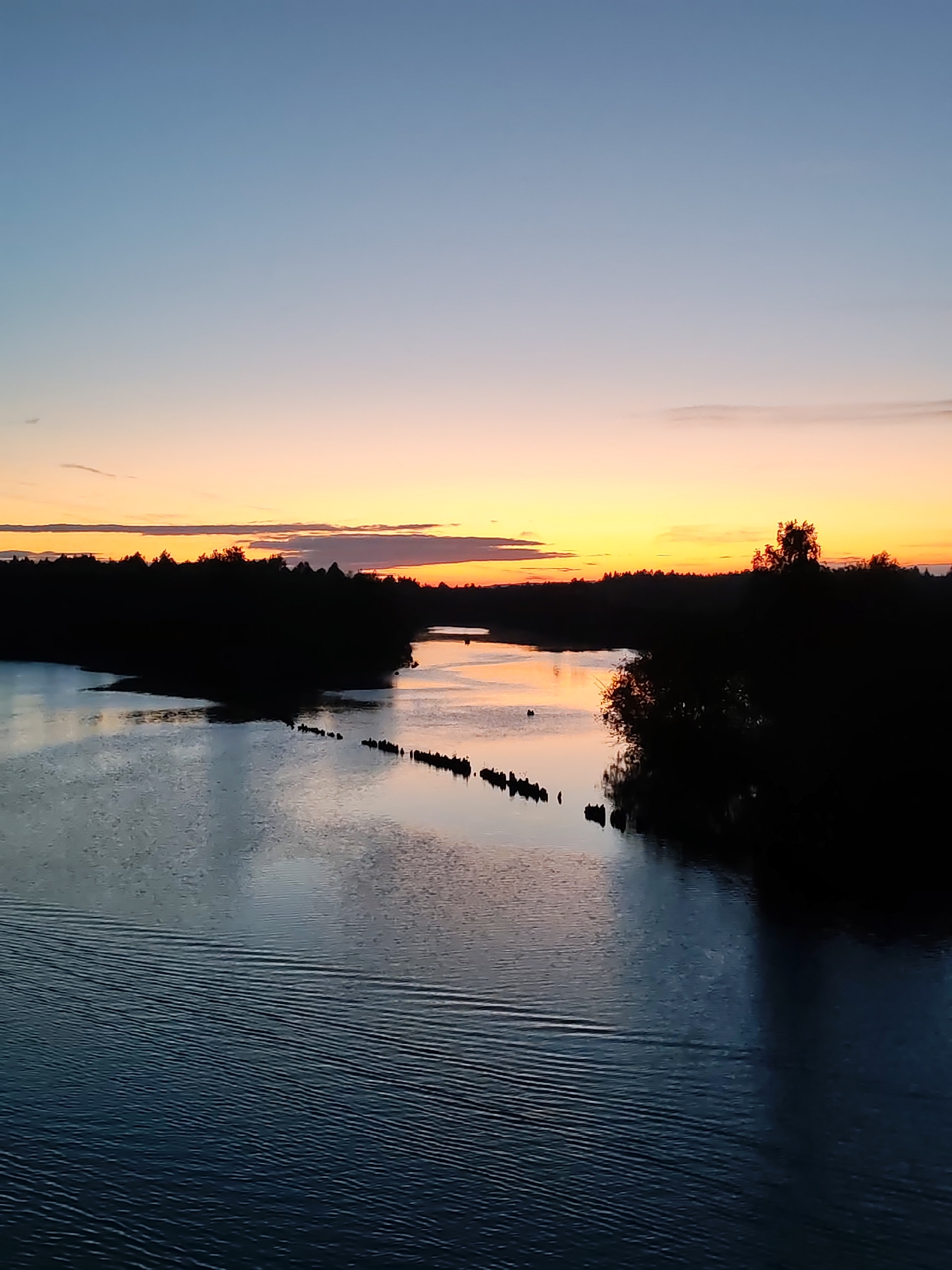 The image captures a sunset view over a river in Annensky Most, Russia. The sky transitions from blue to orange and yellow near the horizon, reflecting in the water. Silhouetted trees line the riverbanks, and remnants of pilings are visible in the river. Ripples are present on the water surface, and the overall scene conveys a sense of tranquility.