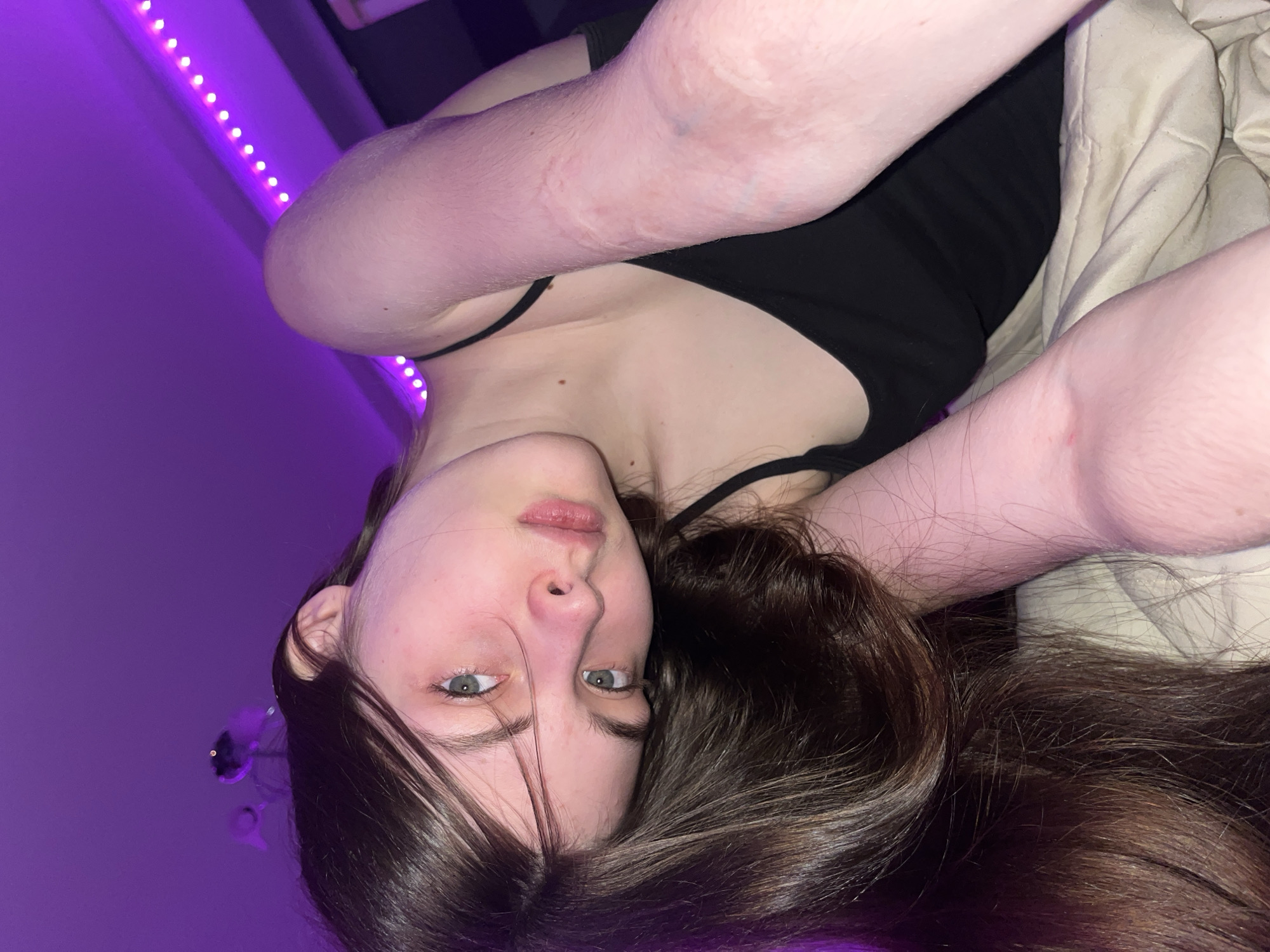A young person with long, dark hair and fair skin is lying down, possibly on a bed covered with a light-colored comforter. They are wearing a black tank top. The wall behind them is lit with purple LED lights. The background includes a partially visible decorative item on the wall. The location is not discernible as Saint Petersburg, Russia.