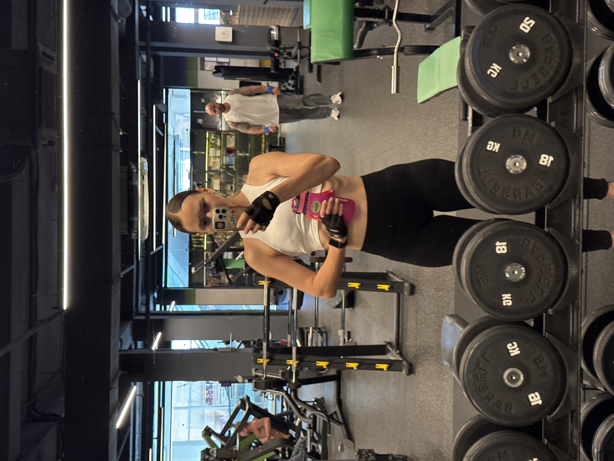 In a gym located in Sochi, Russia, a woman with dark hair tied back, wearing a white tank top and black leggings, stands facing a mirror. She holds a pink water bottle in her left hand and is taking a selfie with her phone. In the background, a man with a shaved head and a white tank top is visible, walking away from the camera. The gym is equipped with various weightlifting machines and a rack of dumbbells.
