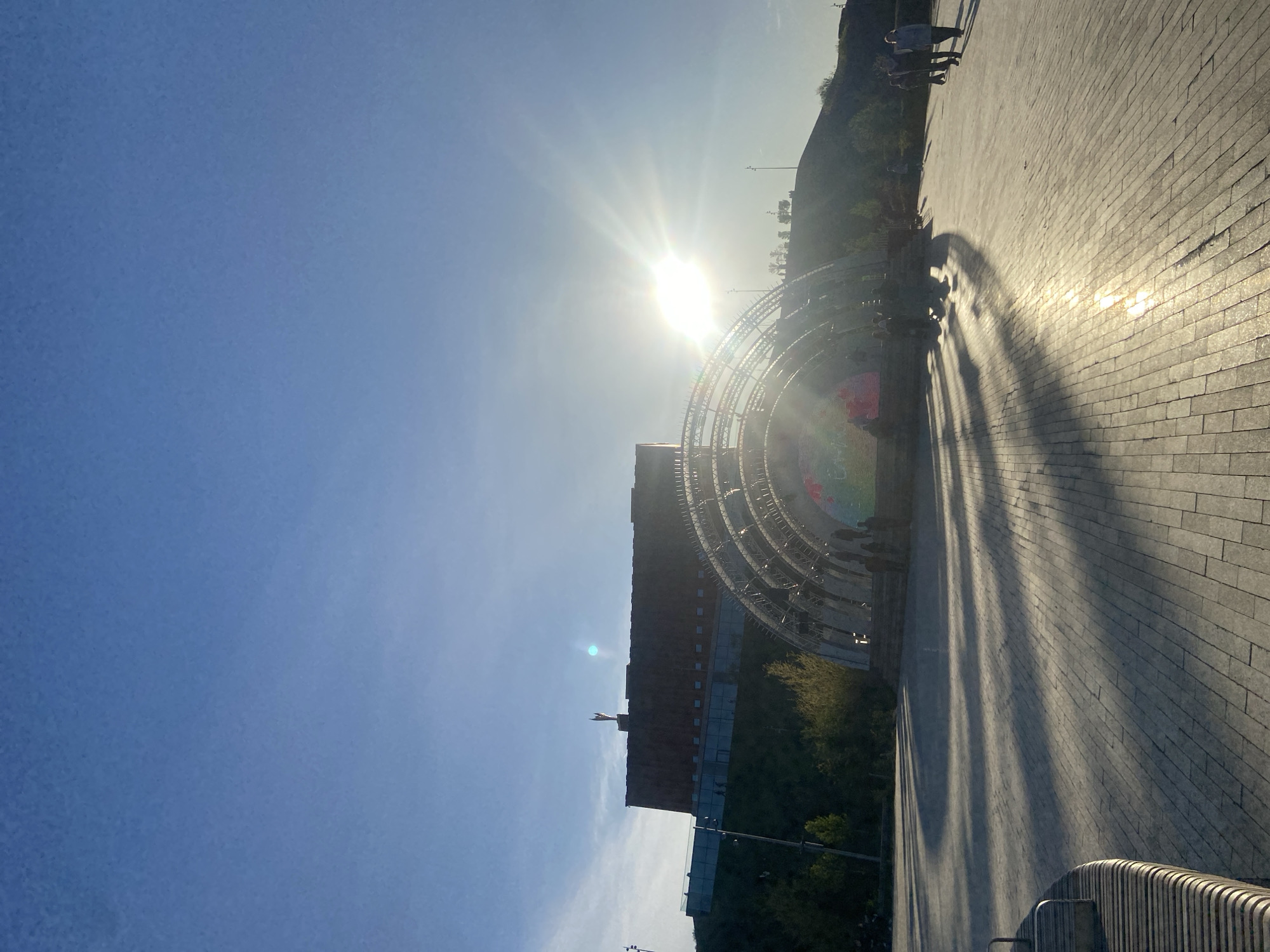The image depicts a view of Kazan, Russia, under bright sunlight. The city's architecture is visible, with a tall brick building topped with a spire. A large circular structure with concentric rings is visible in front of the building. People are walking on a wide, paved walkway that stretches to the right of the frame. Shadows from the circular structure fall across the walkway. The sky is a gradient of blue, transitioning from a darker shade to a lighter one closer to the sun. There is foliage to the right, appearing as a green hill, potentially an embankment or park area.