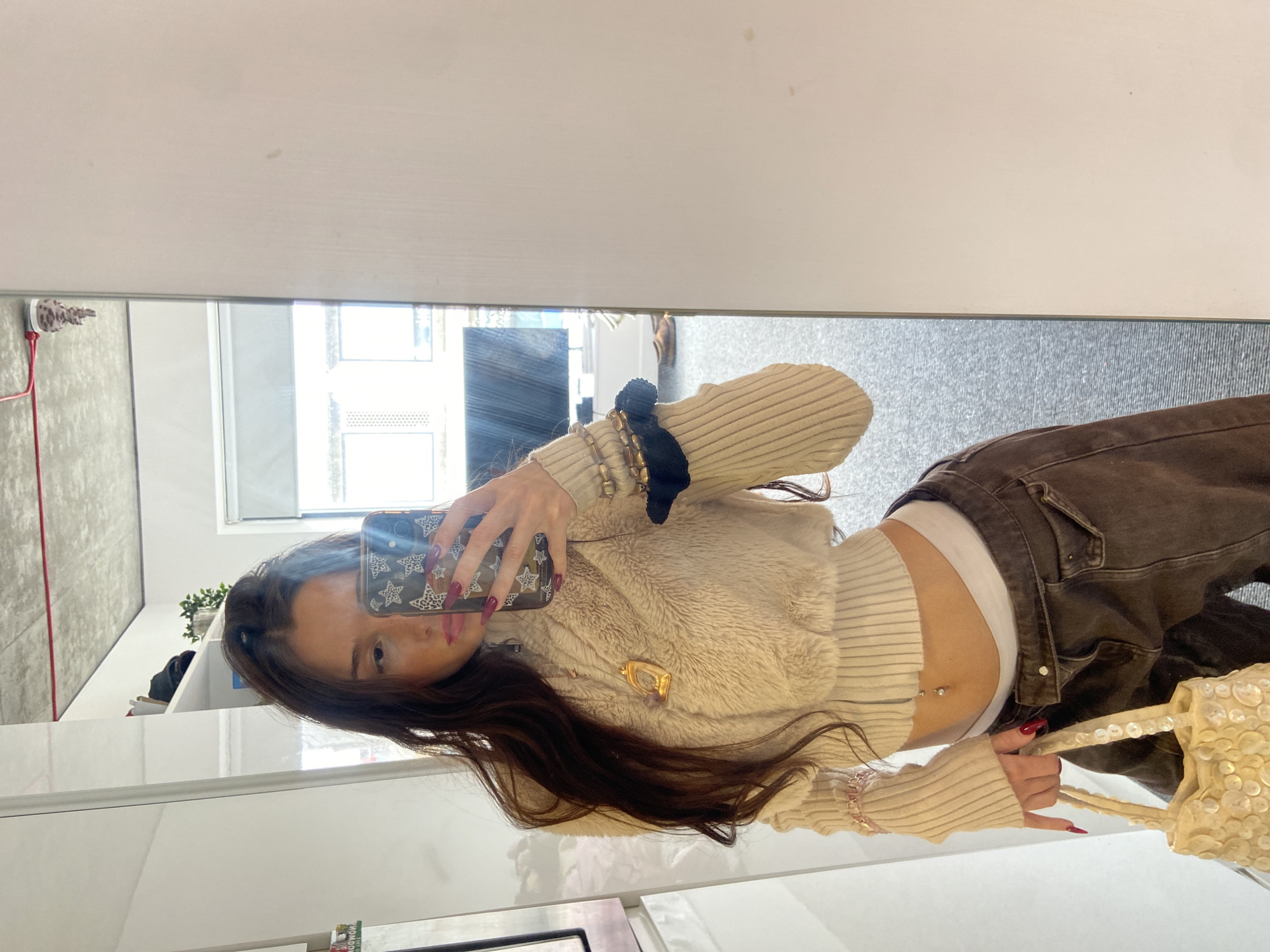 This mirror selfie, rotated sideways, captures a young woman posing indoors, likely in an apartment or modern room in London, United Kingdom. She looks directly into the camera with a neutral expression.
The woman has long, dark brown hair and is dressed in a layered outfit. She wears a light beige or cream ribbed long-sleeved sweater, topped with a fluffy or faux fur vest of a similar color. A white undershirt is visible at her waist, complementing her dark brown or grey jeans.
Her accessories are prominent: multiple bracelets and a black scrunchie adorn her left wrist, while a distinctive gold-toned brooch with a purple inlay is pinned to her vest. Her fingers are painted with bright red nail polish, and she has a silver barbell piercing on her navel. She holds her phone, encased in a black cover with white star patterns, to take the picture, while her right hand grasps a small, light-colored, sequined or embellished handbag.
The background, reflected in the mirror, shows a contemporary interior. To the left, a grey, possibly concrete or textured, wall is visible. A large window provides natural light, suggesting it is daytime, and offers a glimpse of an outdoor scene, possibly other buildings. White shelving with some small green plants can be seen, along with a grey carpet or floor covering. No explicit text is visible in the image.