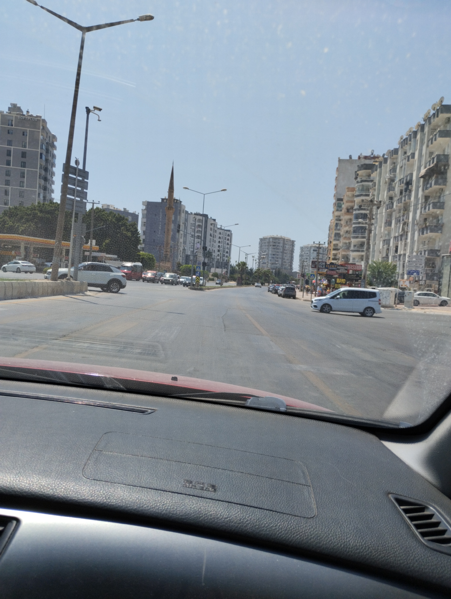 The image captures a street scene in Mezitli, Turkey, as seen from the interior of a vehicle. The foreground shows the dashboard of a car, with the windshield obscuring the view ahead. A red-colored dashboard is partially visible. Beyond the windshield, a wide, paved road stretches into the distance, with multiple lanes of traffic. Various vehicles, including cars and SUVs, are present on the road.
On either side of the road, large, multi-story apartment buildings line the street, characteristic of urban architecture. These buildings feature numerous balconies and windows. A prominent minaret of a mosque is visible among the buildings on the right side of the road, indicating the presence of a religious site. Streetlights and utility poles are also visible along the roadside. The sky is clear and blue, suggesting a sunny day. In the distance, trees and other urban structures contribute to the city landscape. The overall impression is a typical daytime view of a busy street in a Turkish coastal city.